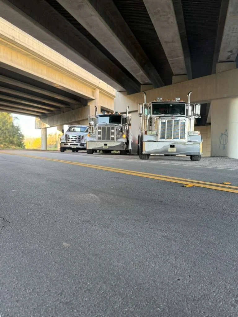 Two large silver trucks and one silver car parked under an overpass on a road.