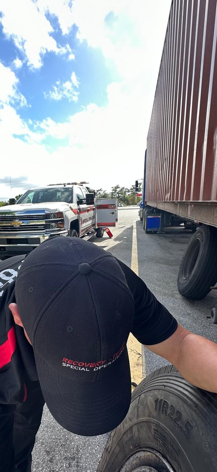 A recovery worker adjusting equipment next to a large truck and a police vehicle in a parking lot on a partly cloudy day.