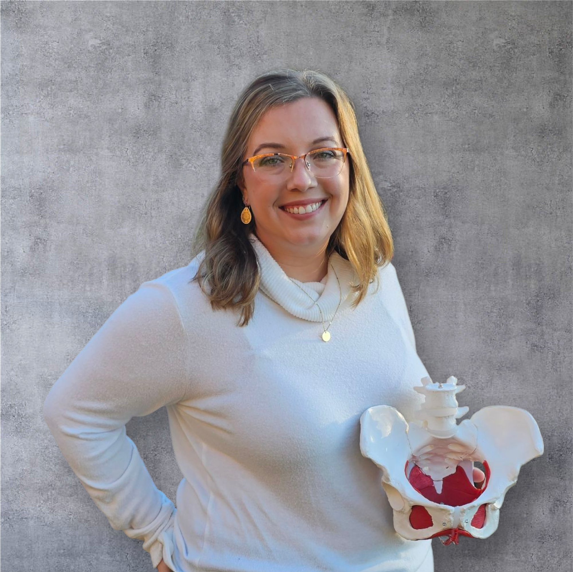 A woman wearing glasses and earrings, holding a model of a human pelvis, standing against a gray concrete wall and smiling.