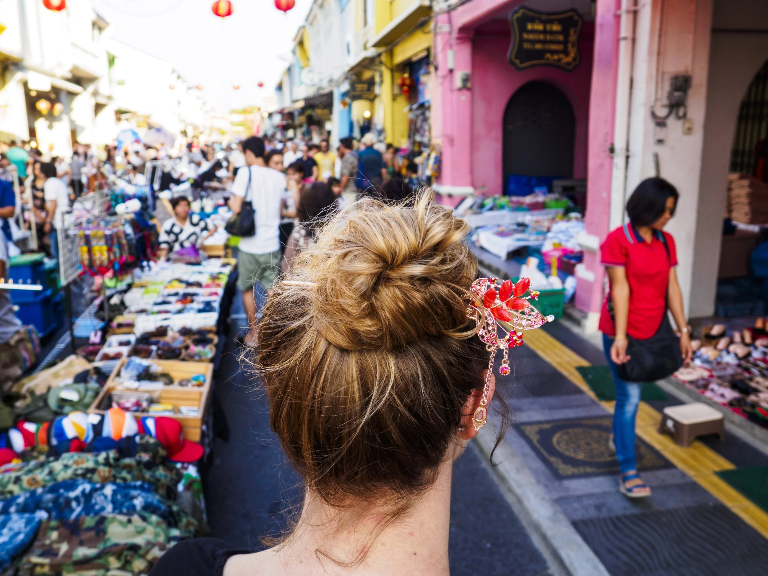 Close-up of a woman's head with styled hair and decorative hair accessory, she is at a crowded street market with stalls selling various items, colorful buildings, and shoppers in the background.