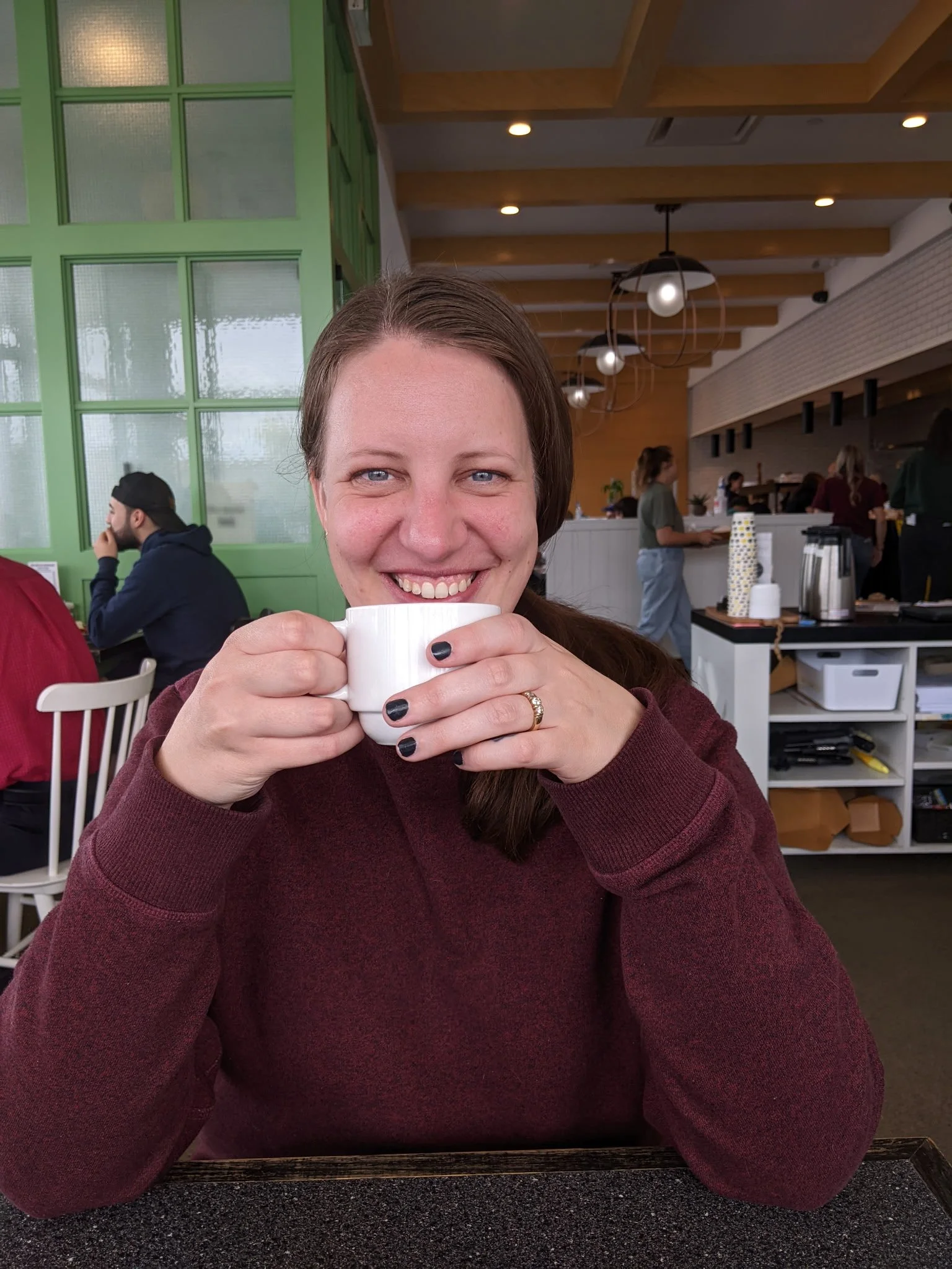 A woman with long brown hair and a maroon sweater sitting at a table holding a white mug, smiling at the camera in a modern cafe with green and white decor and warm lighting.