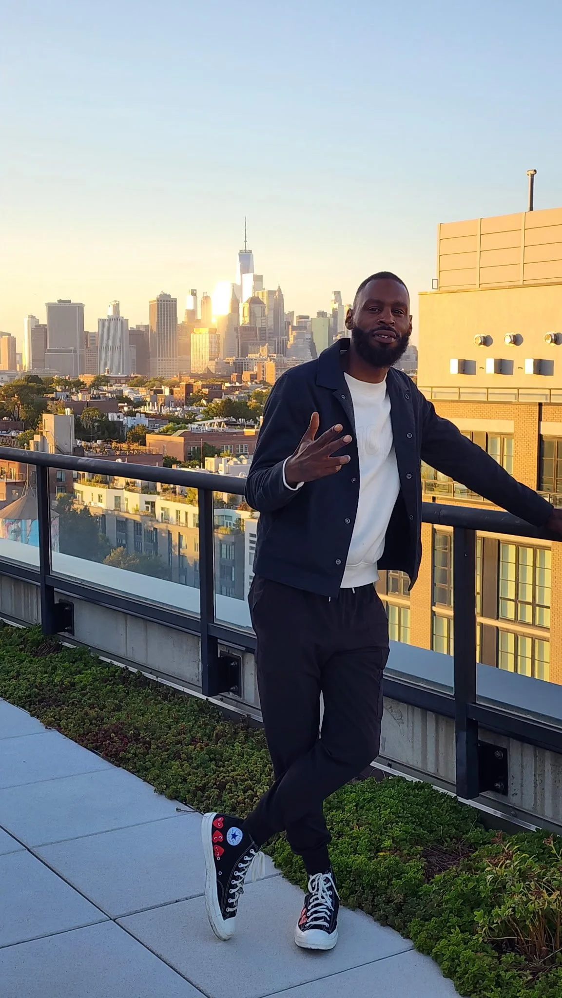 A man standing on a rooftop balcony with New York City skyline in the background during sunset, making a peace sign with his fingers.