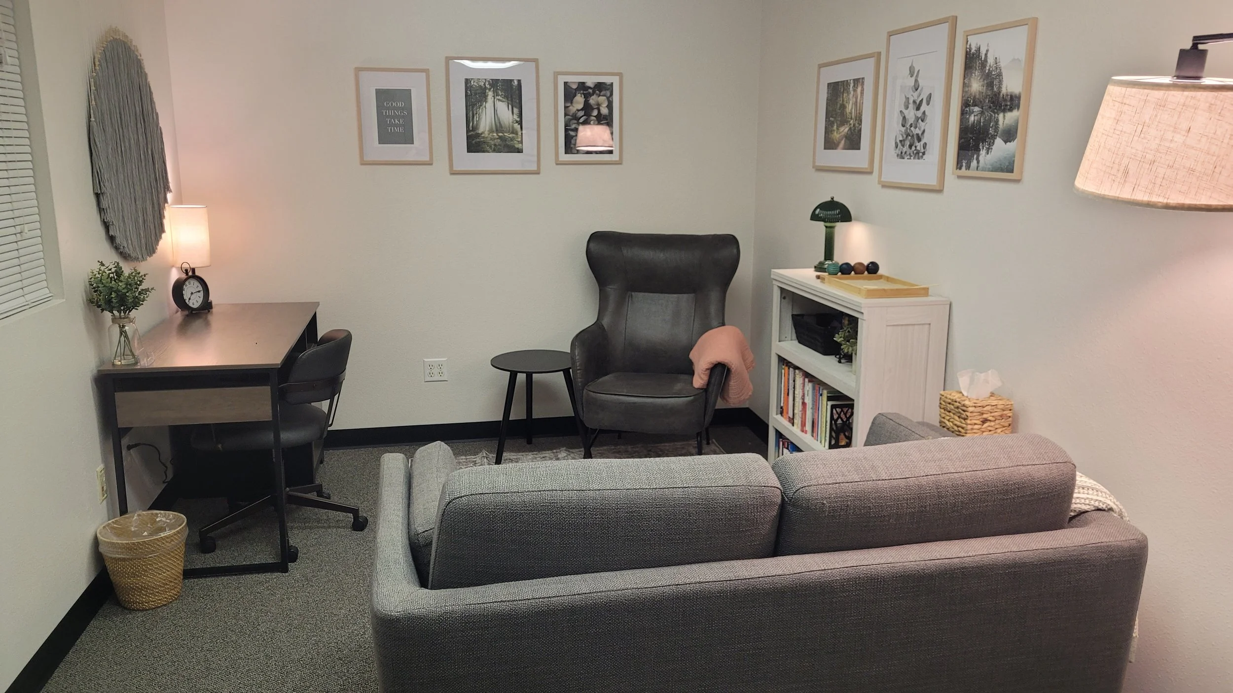 Living room with a gray couch, black leather armchair, small black side table, white bookshelf with decorative items, framed artwork on the wall, desk with a lamp, clock, plant, and chair, carpeted floor, and a window with blinds.