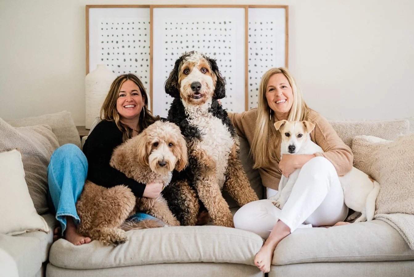 Two women sitting on a beige couch with three dogs in a cozy living room, smiling at the camera.