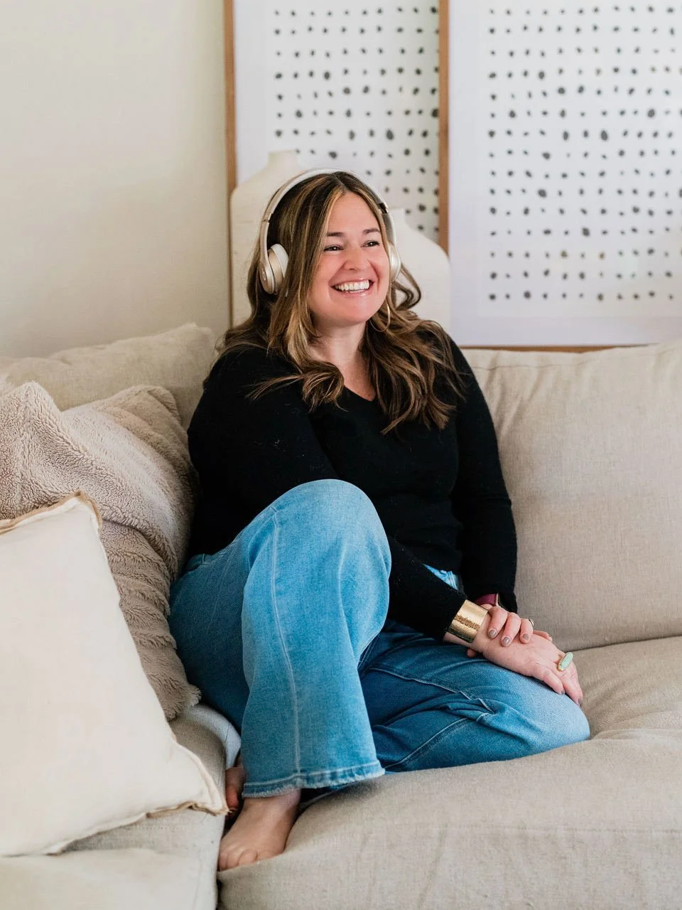 A woman sitting on a beige couch, smiling, wearing headphones, a black top, and blue jeans.