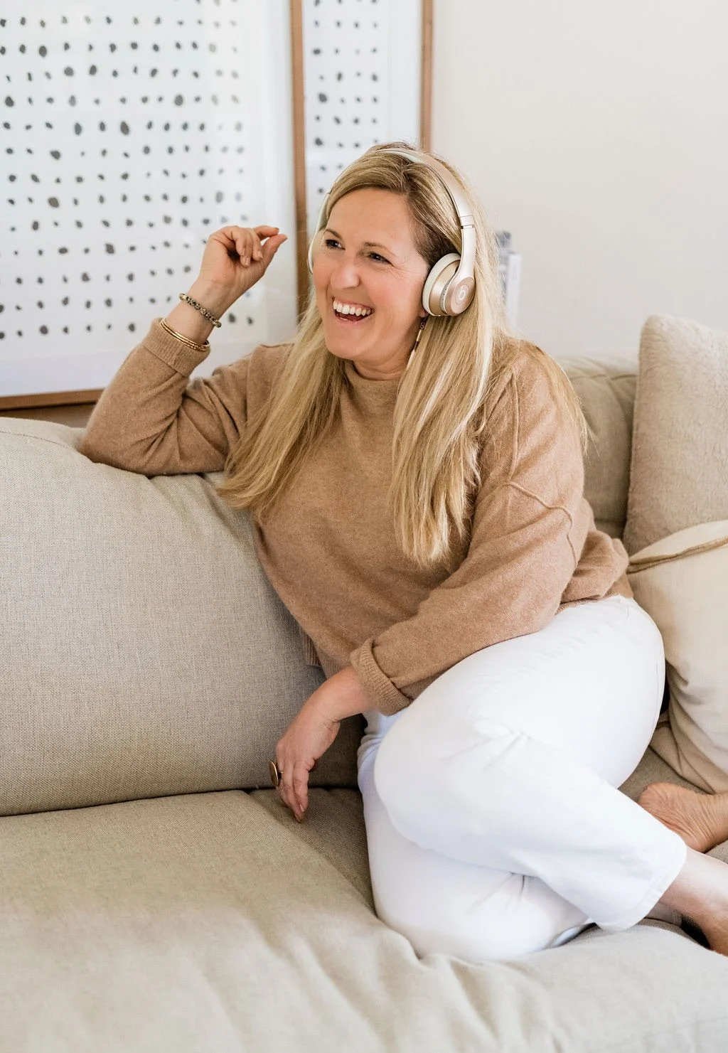 A woman with long blonde hair, wearing a tan sweater and white pants, sitting on a beige couch, smiling and listening to music with over-ear headphones.
