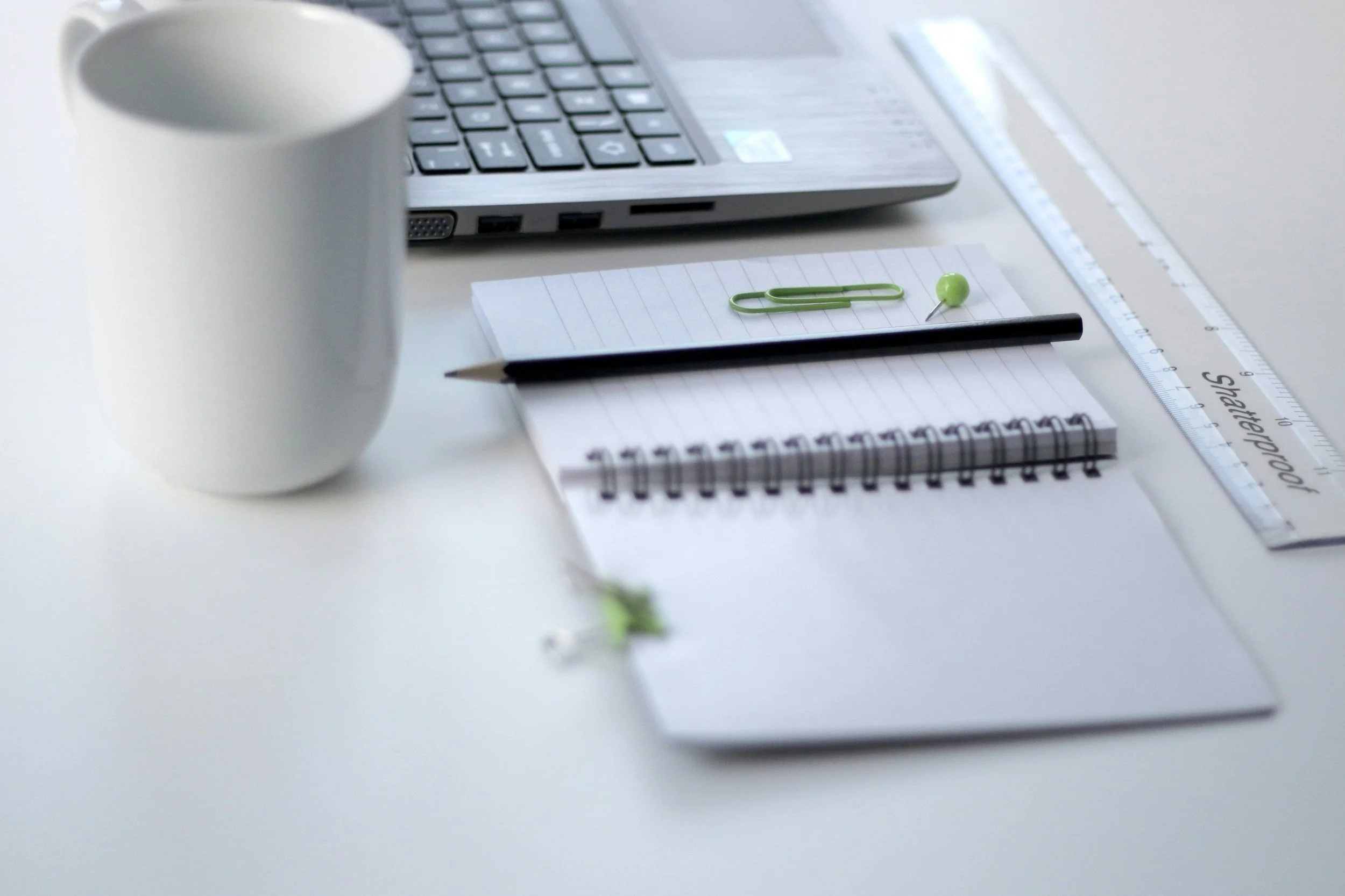 A white desk with a laptop, a spiral notebook, a black pen, a lined notepad, a green paperclip, a green push pin, a transparent ruler, and a white coffee mug.