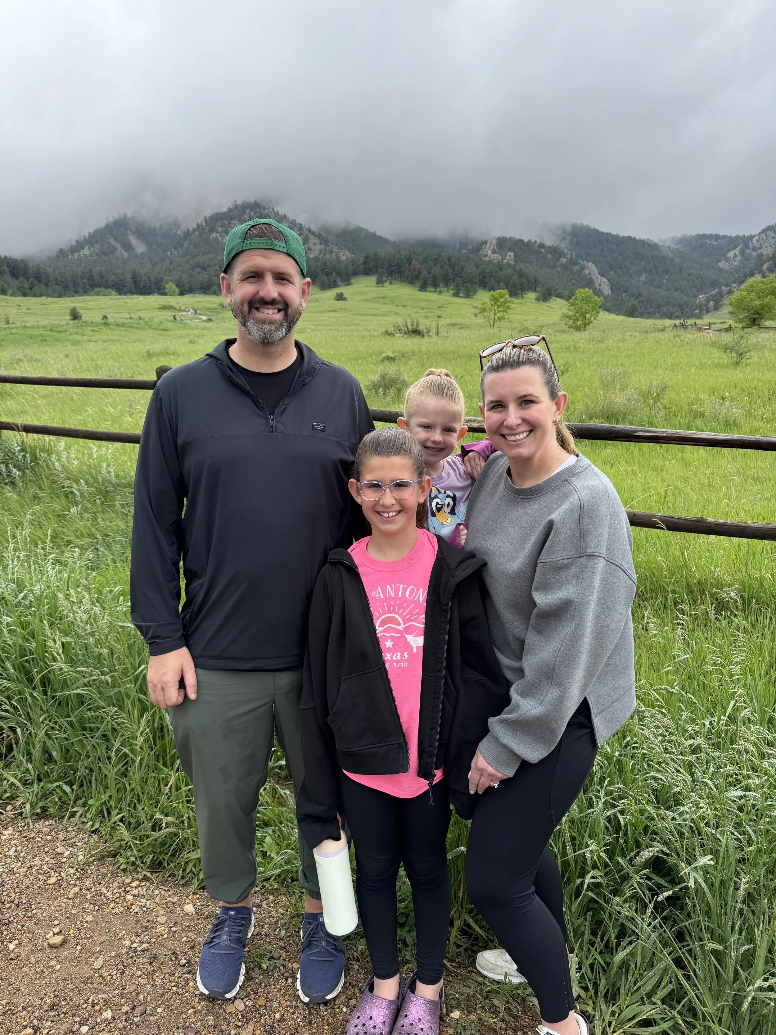 A smiling family of four outdoors on a cloudy day, standing on a dirt path in front of a green field with rolling hills and mountains in the background. The father is wearing a black jacket and green cap, the mother in a gray sweatshirt, the daughter in a pink shirt and black jacket, and the young girl in a light purple dress with a cartoon character.
