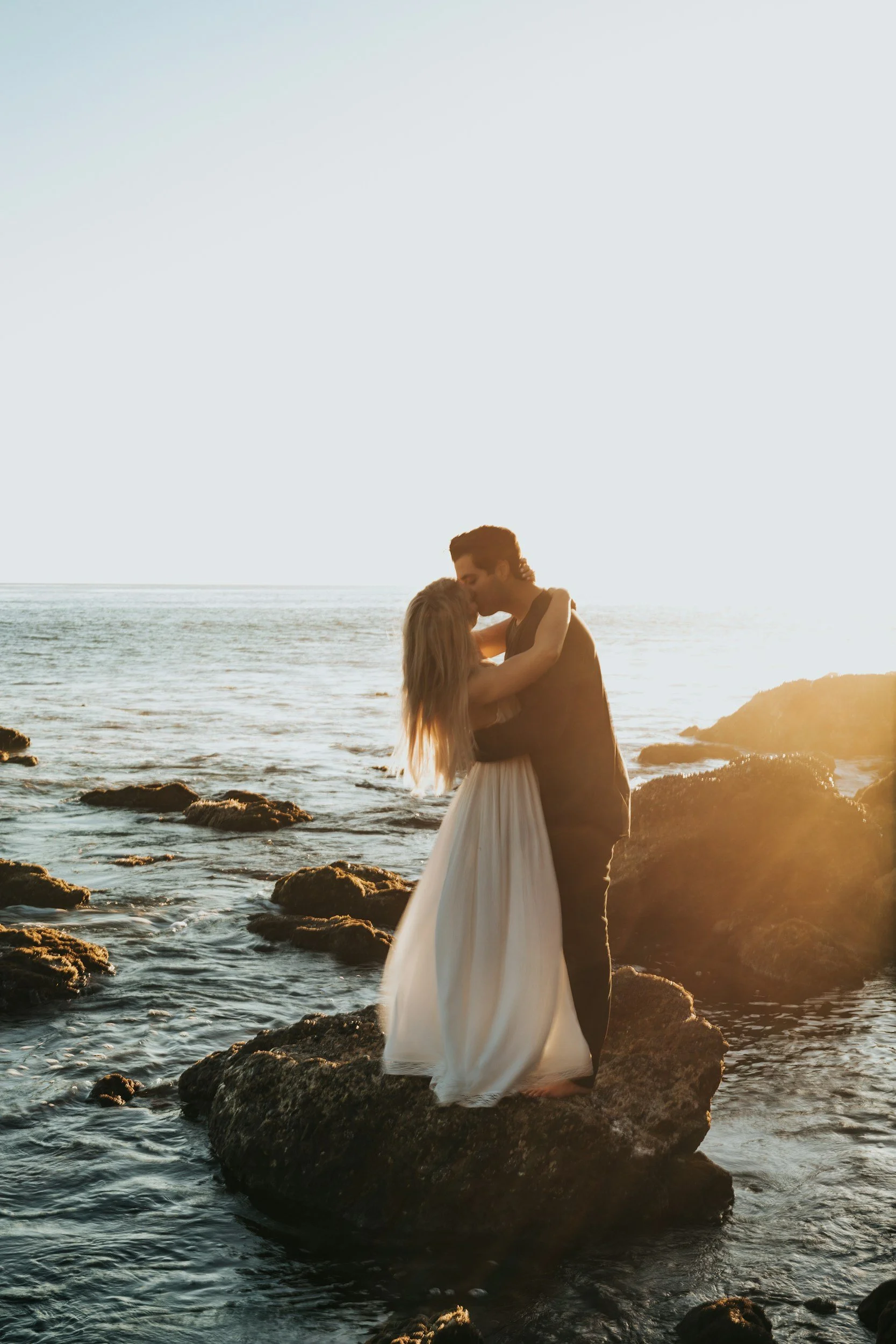 A couple kissing on rocks by the ocean at sunset, with the woman in a white dress and the man in dark clothing.