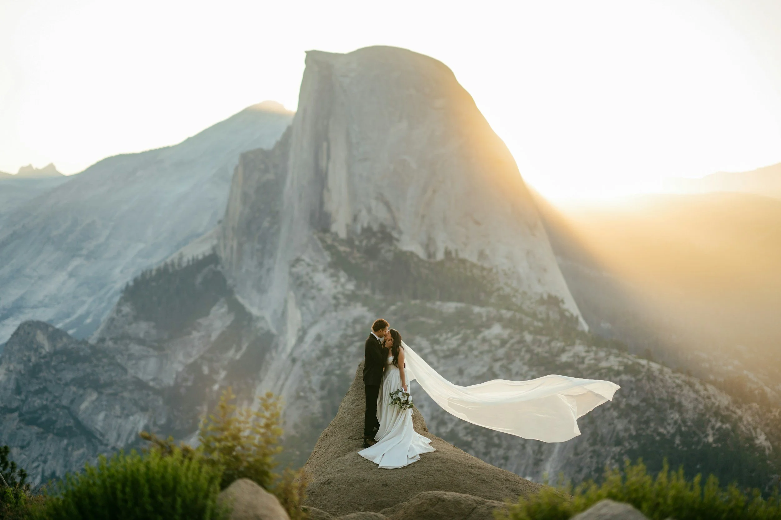 Bride and groom kissing on a rock with Half Dome in the background at sunset.