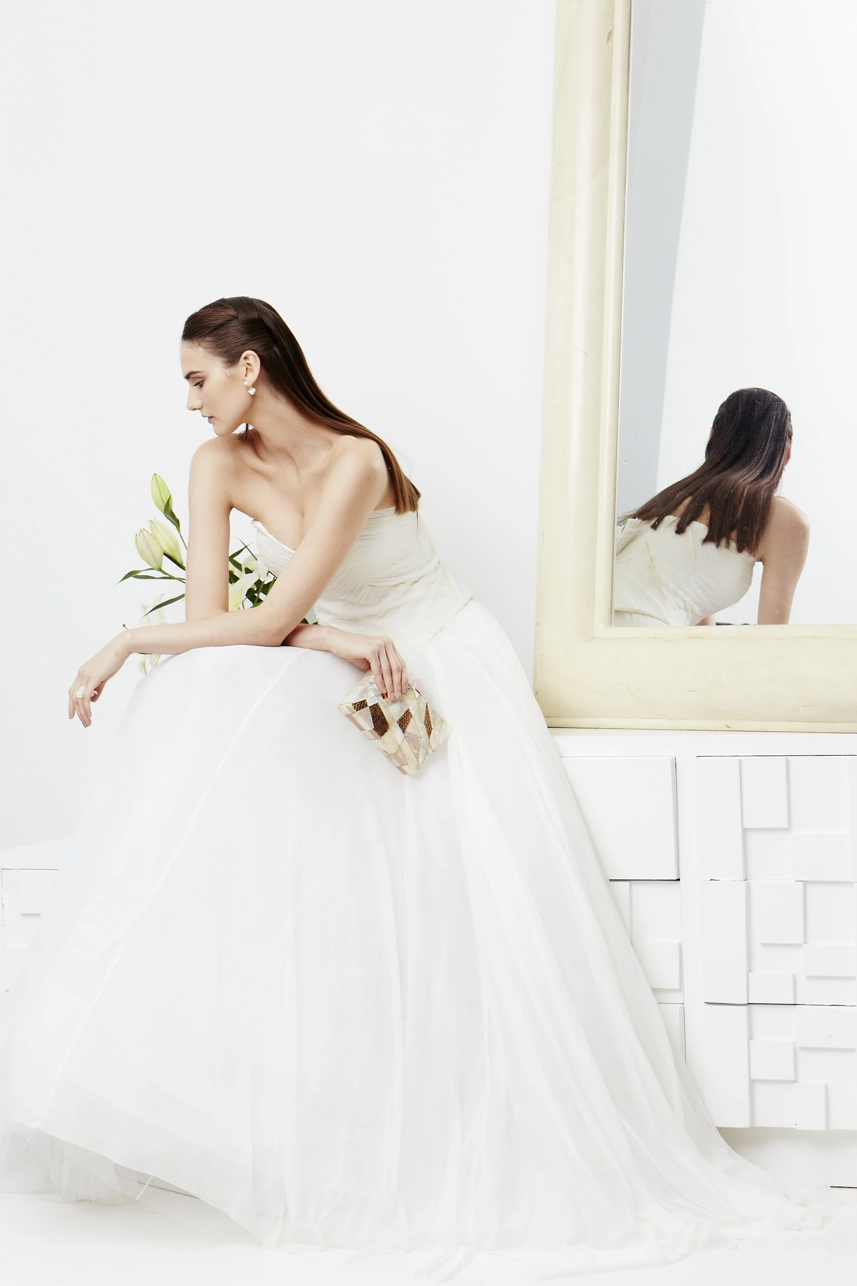 A woman in a white wedding dress leaning on a white surface, holding a clutch bag, with a mirror reflecting her back, in a minimalist white room with a large mirror and flowers.