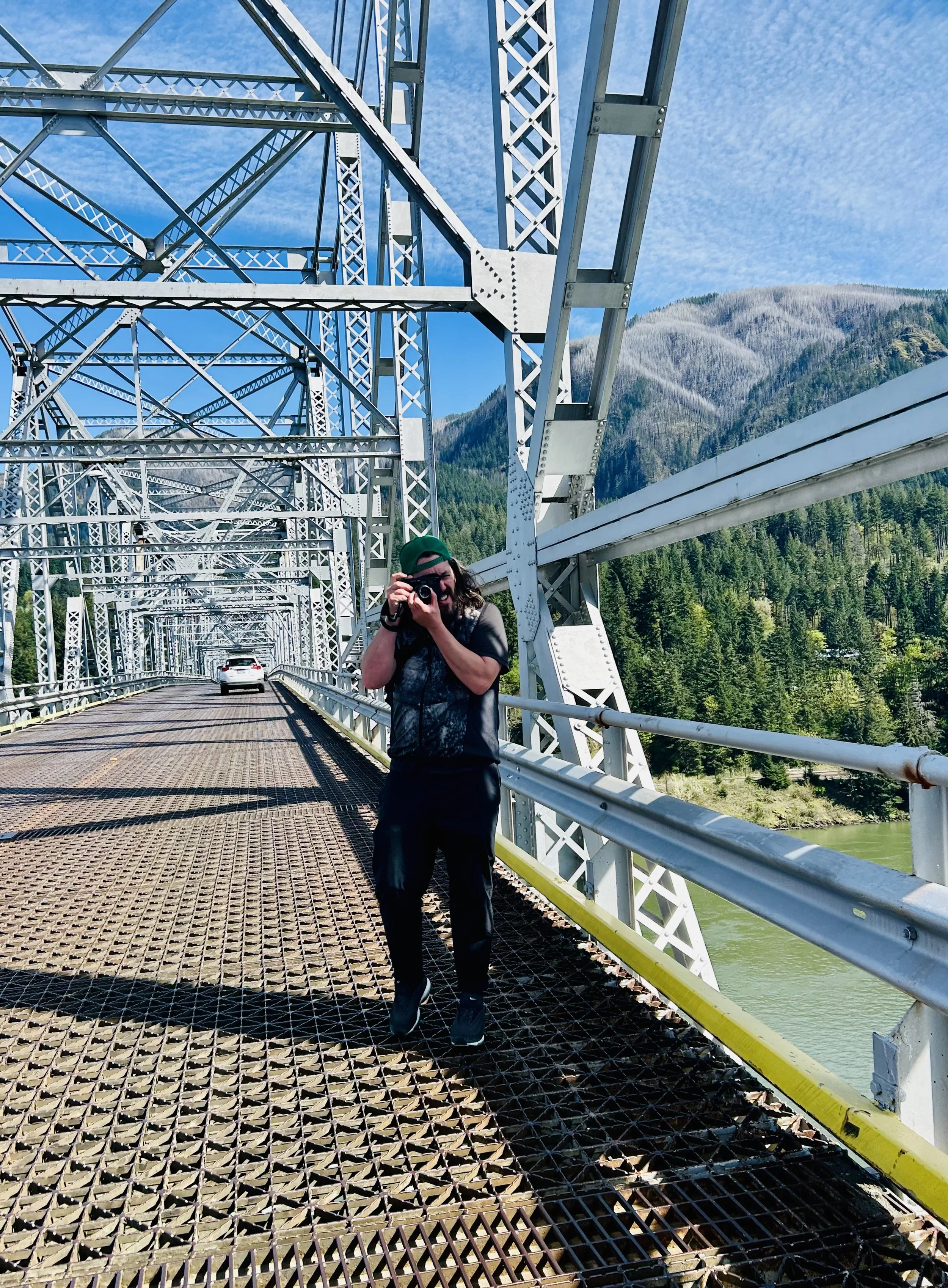 Person taking a photo on a metal bridge with mountains and forest in the background.