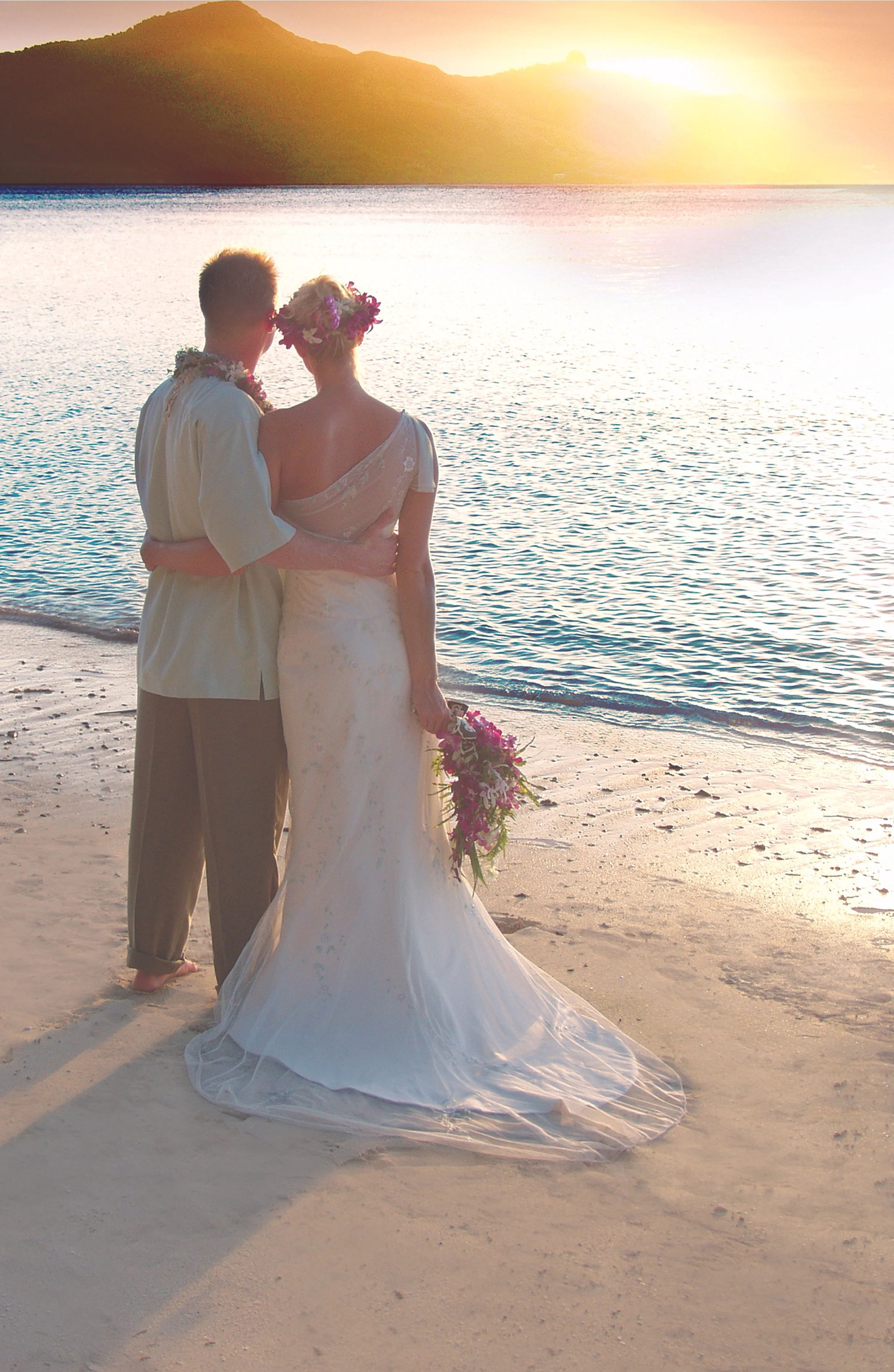 A couple in wedding attire standing on the beach at sunset, overlooking the ocean with a mountain in the background, holding a bouquet, embracing each other.