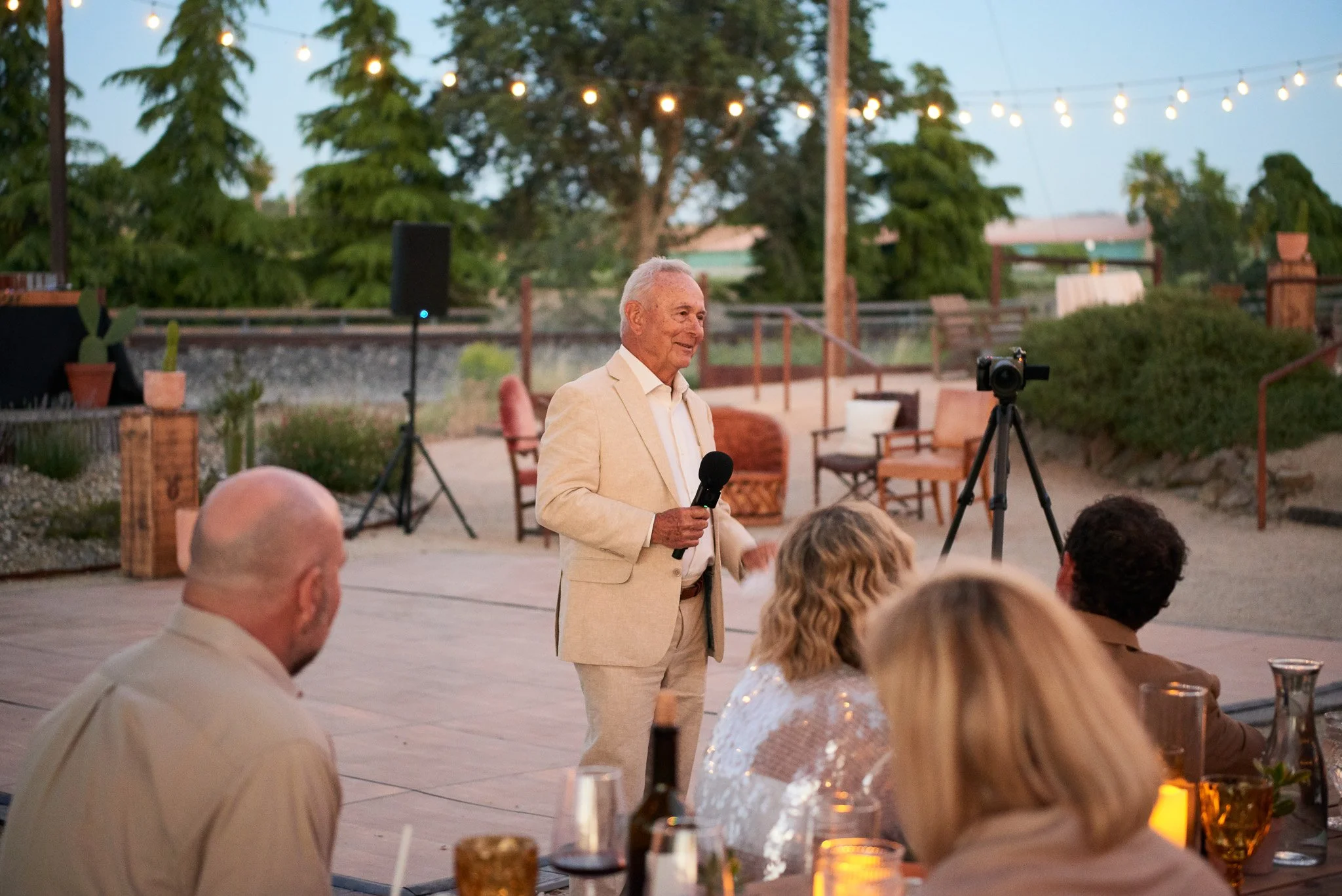 An elderly man in a beige suit giving a speech with a microphone at an outdoor evening event, with guests seated at a table in the foreground, string lights overhead, and a camera on a tripod capturing the moment.