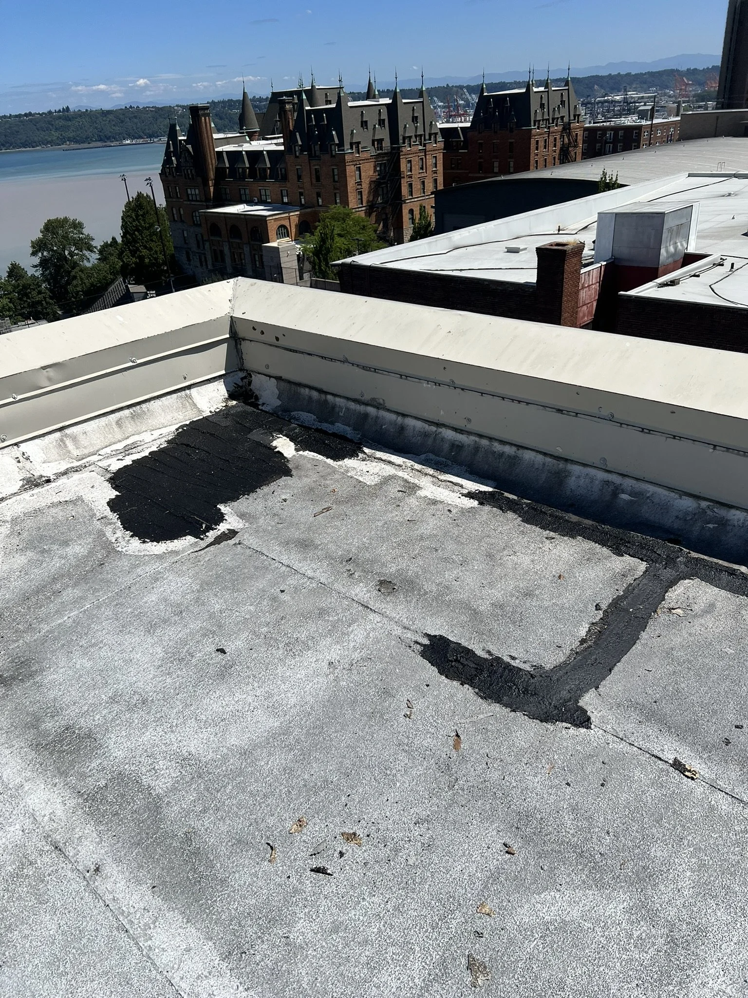 View of a rooftop with a worn flat surface, some patchwork repairs, and a white metal parapet against a cityscape background with historic buildings, trees, and distant hills under a partly cloudy sky.