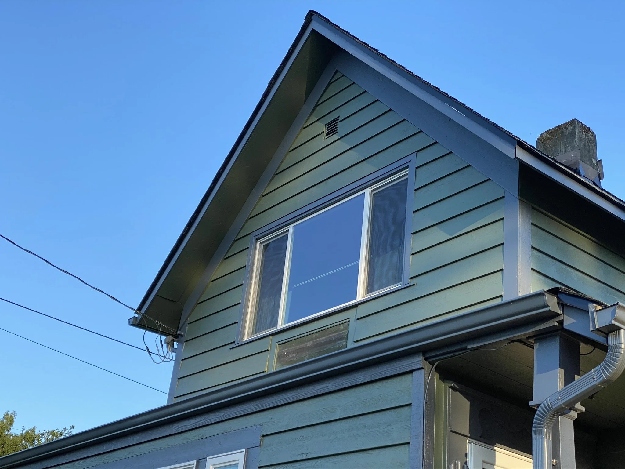Close-up of a light green house with horizontal siding, a large window, and a steep gabled roof against a clear blue sky.