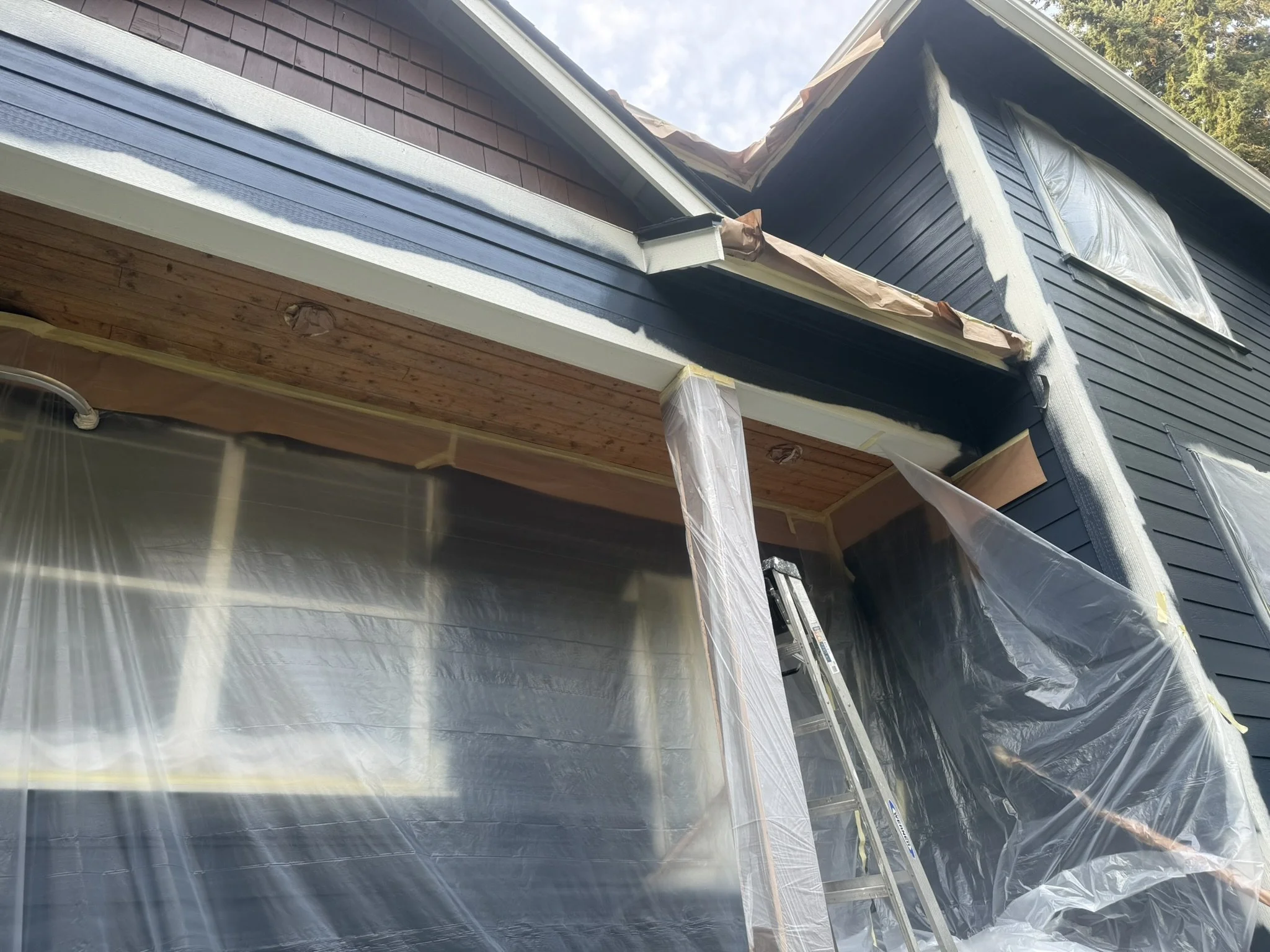 House undergoing exterior renovation, with plastic sheeting covering windows and doors, a ladder leaning against the wall, and new soffit installation visible under the eaves.