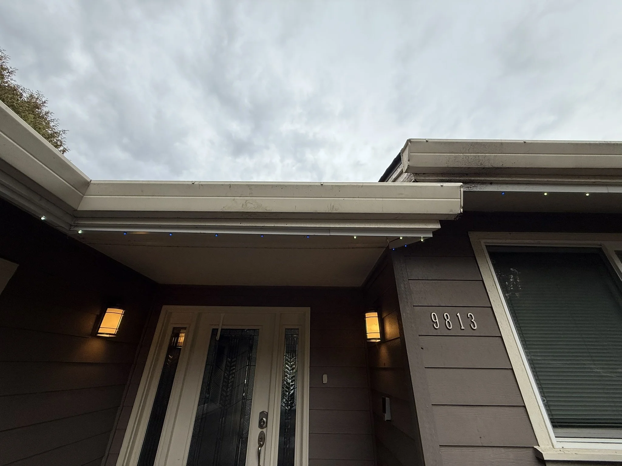 Close-up of house exterior with brown siding, front door, and house number 9813, under cloudy sky.
