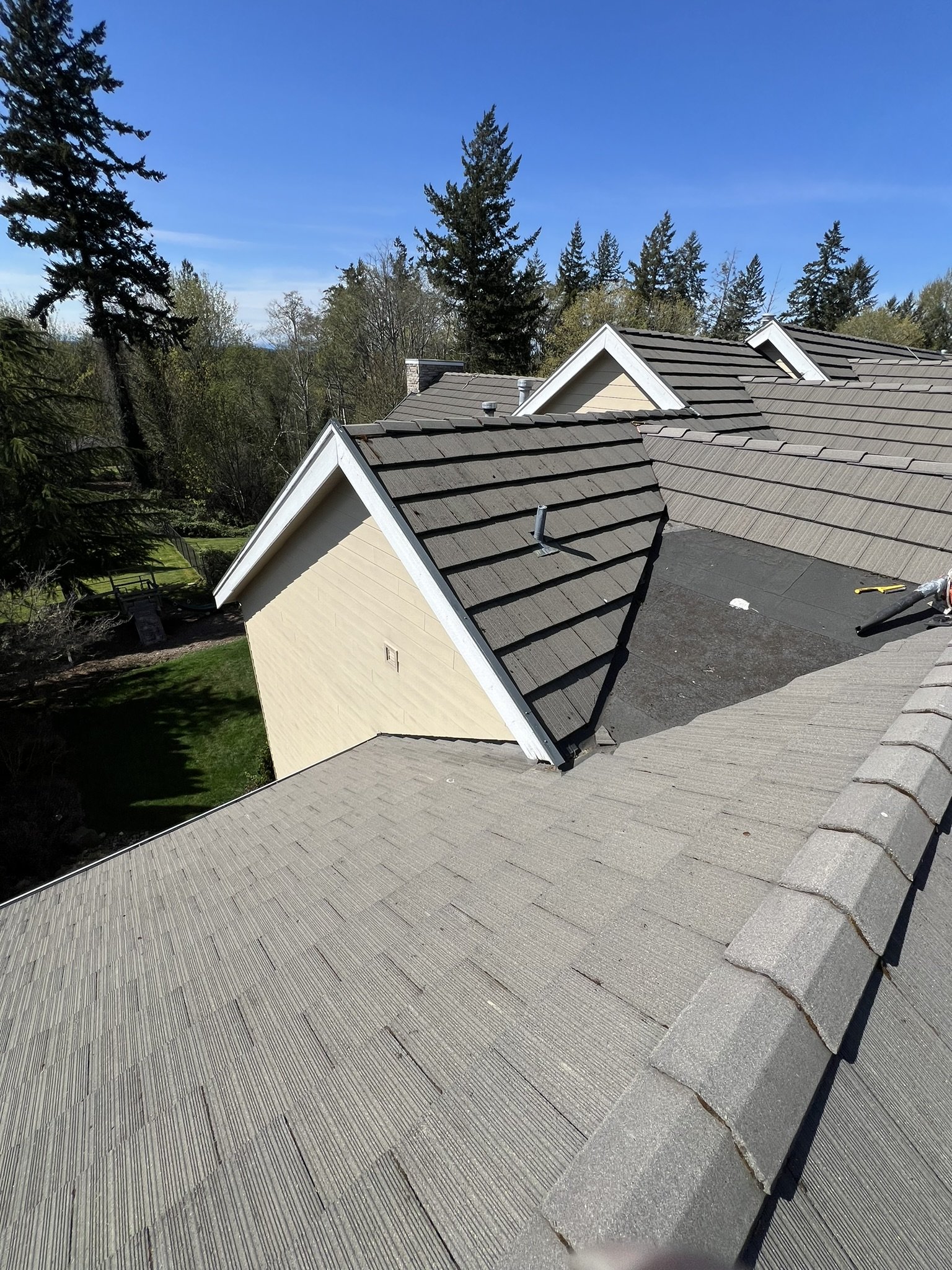 View of rooftops on a residential house under a clear blue sky, with trees in the background. The roof has various sections, some with shingles and one with black underlayment, and construction tools are visible.