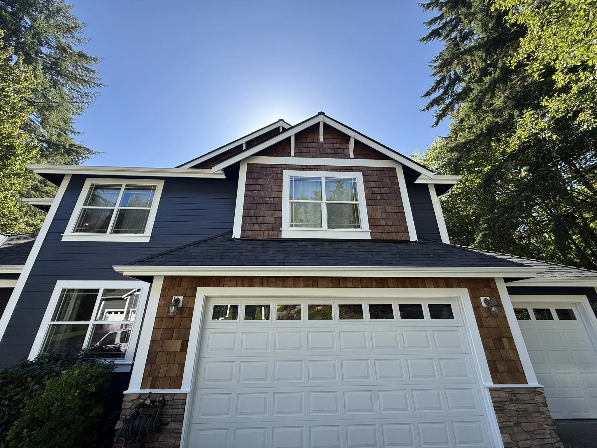 Front view of a two-story house with dark blue siding, brown shingles, and white trim, surrounded by trees under a clear blue sky.