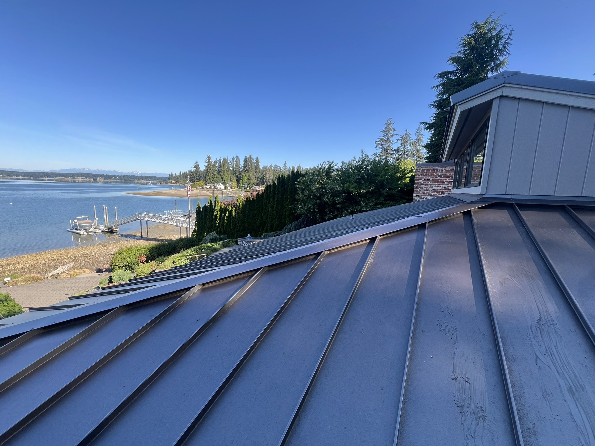 View from a rooftop overlooking a body of water with a small dock, boat, and boat lift. The scene includes green trees, shrubs, and a clear blue sky.