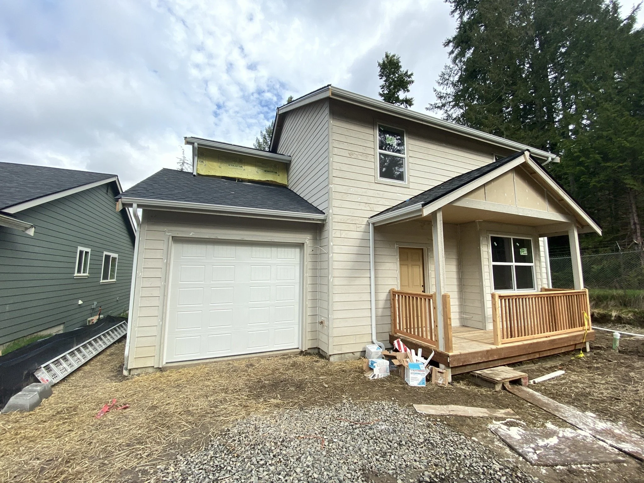 A two-story house under construction with a small front porch, a garage, and beige siding, surrounded by trees and construction materials on the ground.