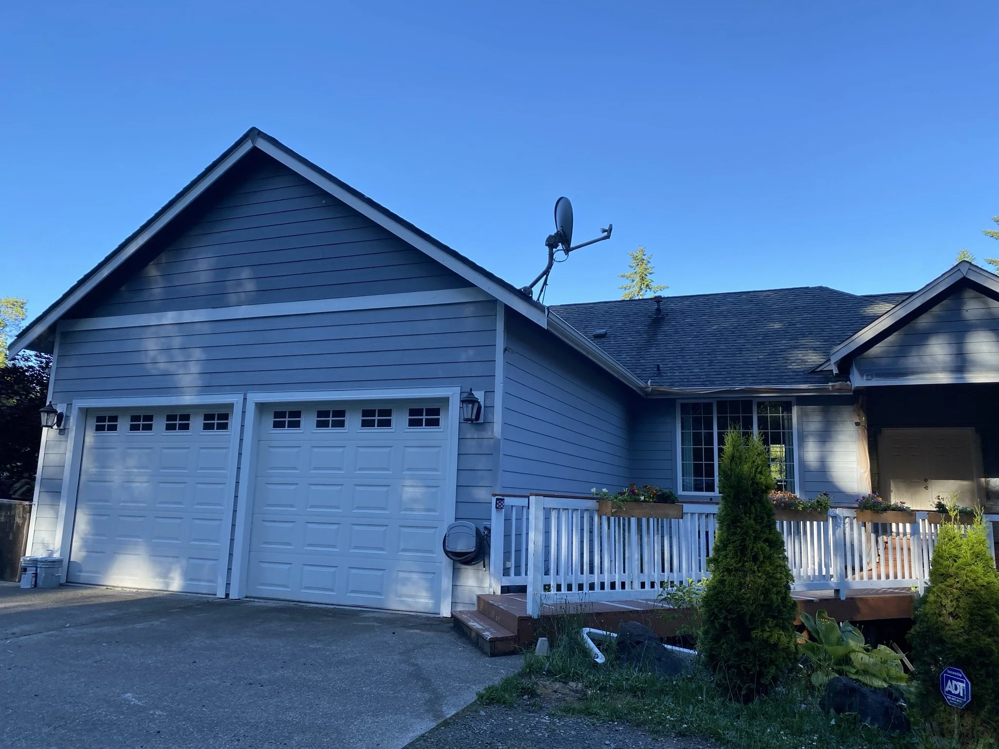Front view of a house with a double garage, blue siding, and a wooden deck with flower boxes. A satellite dish is mounted on the roof, and there are trees and plants in the yard.