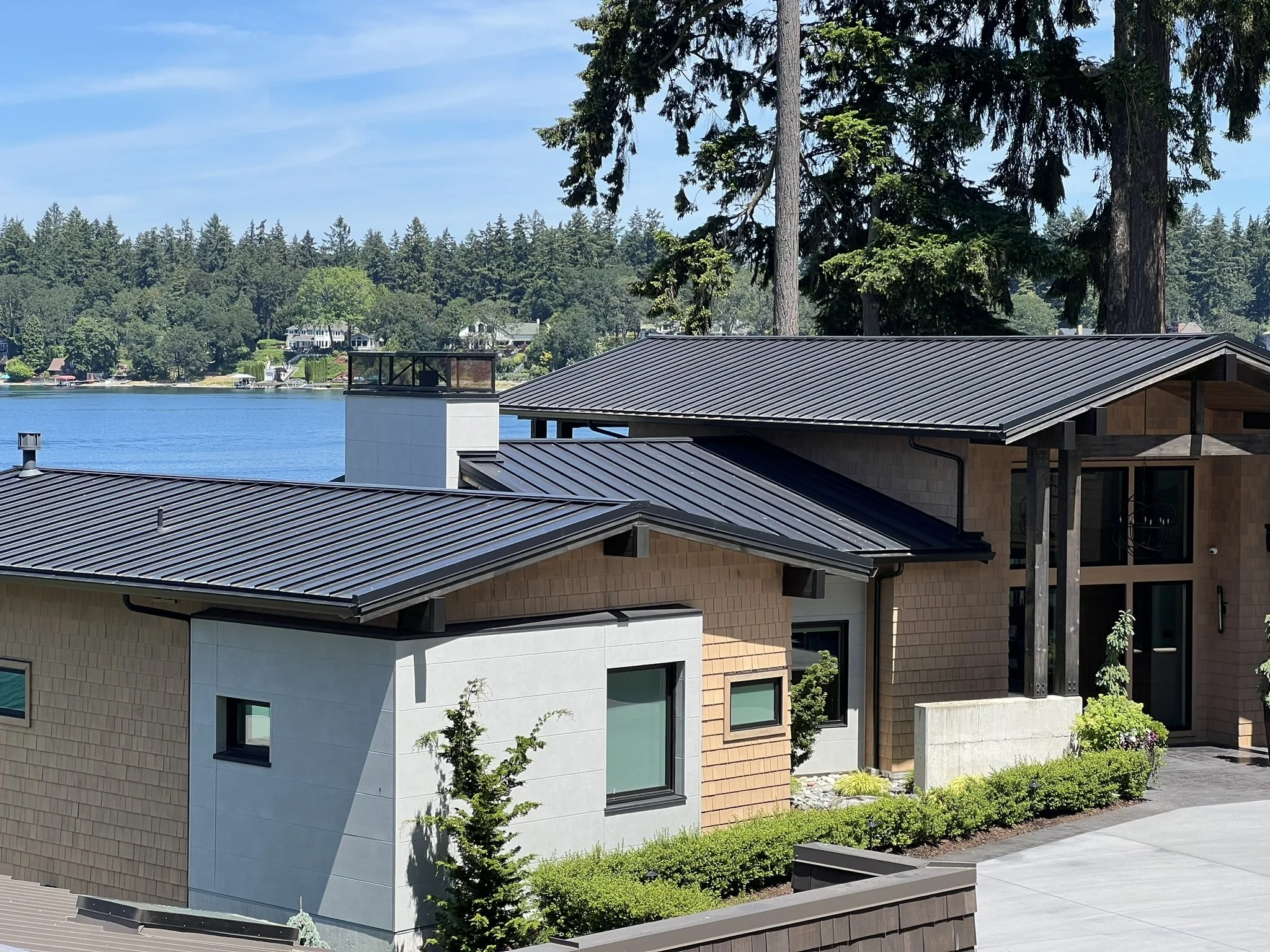 Modern house with black metal roofs, side and front views, surrounded by green shrubs, with trees and a lake in the background.