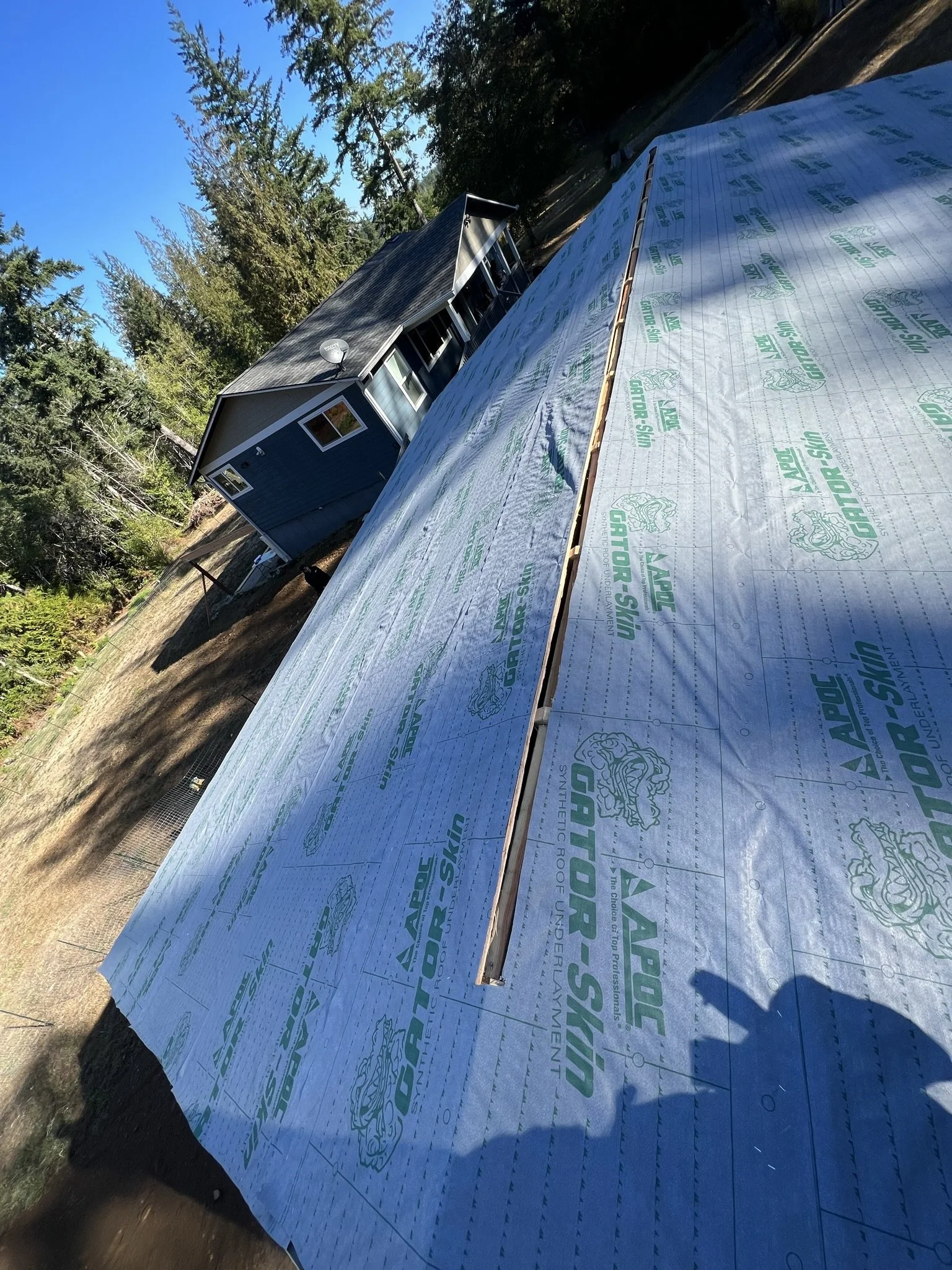 A house under construction with a new roof being installed, covered with a reflective underlayment labeled 'GATOR-SKIN' and 'APEX'. The house is blue, with white trim, and is surrounded by trees.