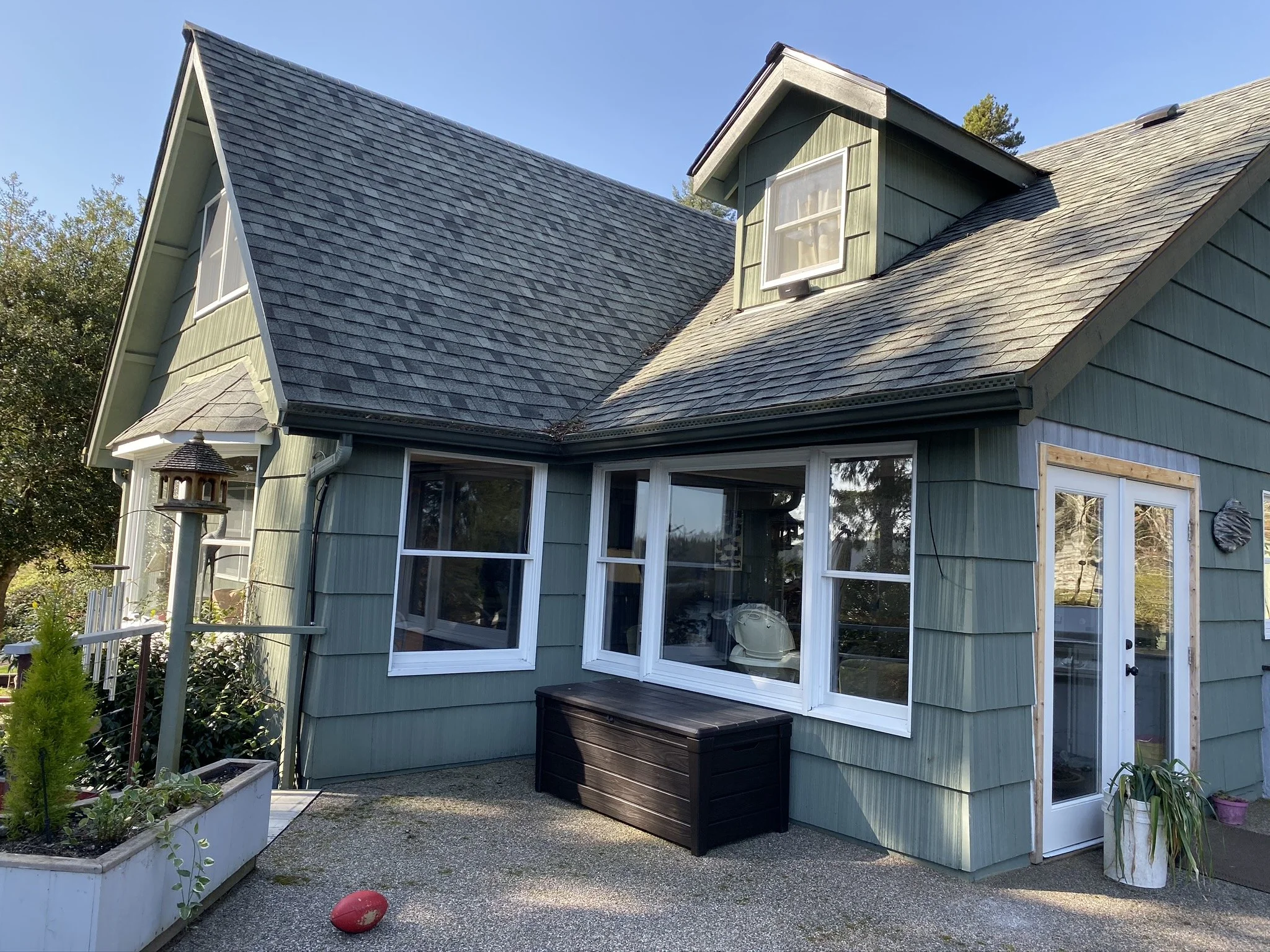 Exterior view of a green house with a gray shingled roof and multiple windows, some with white trims, a small dormer window, a black storage bench, and potted plants on a concrete patio.