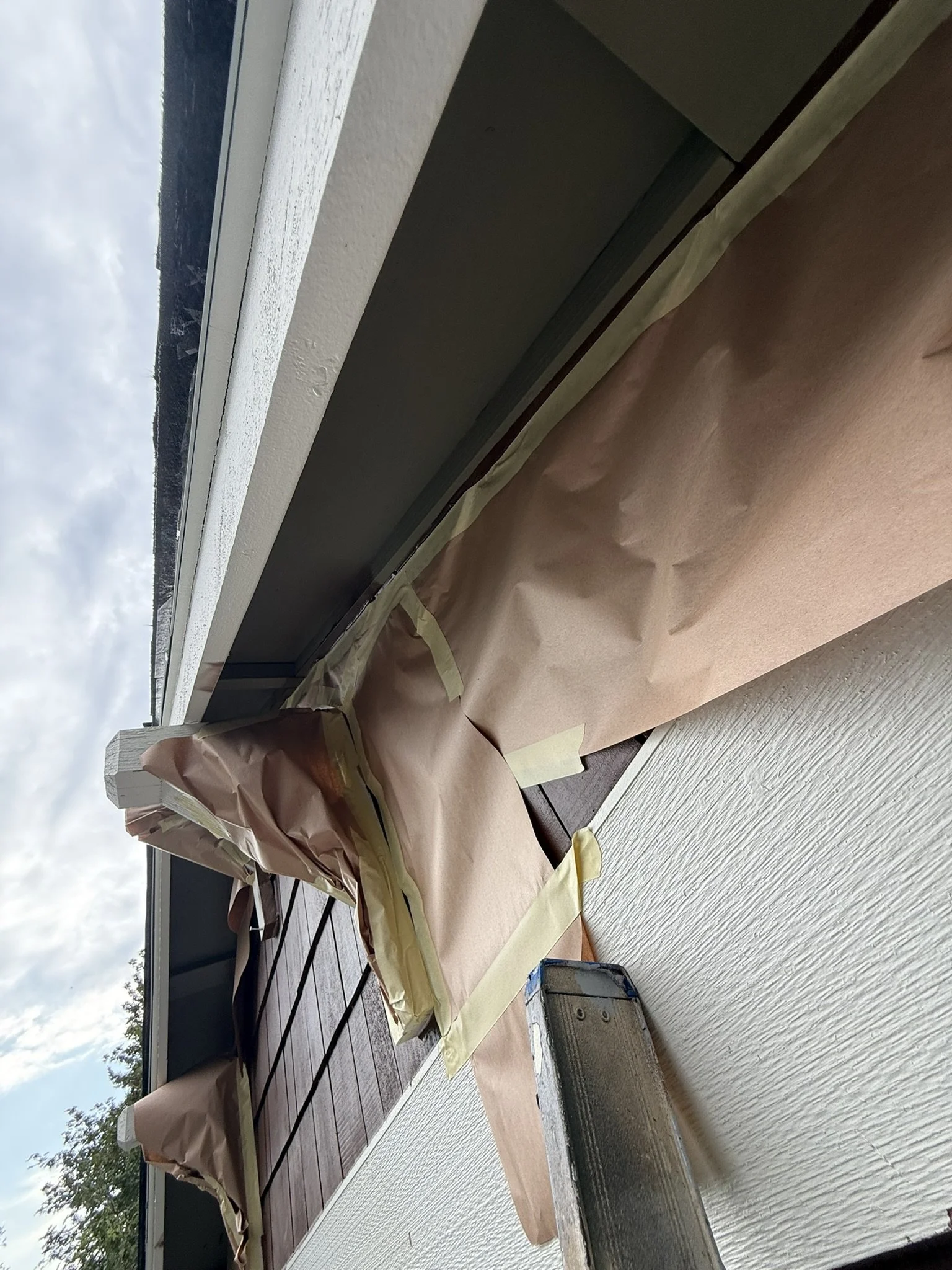 A person applying weatherproofing tape to a home’s balcony ceiling with brown paper masking and tape, with a view of cloudy sky and trees outside.