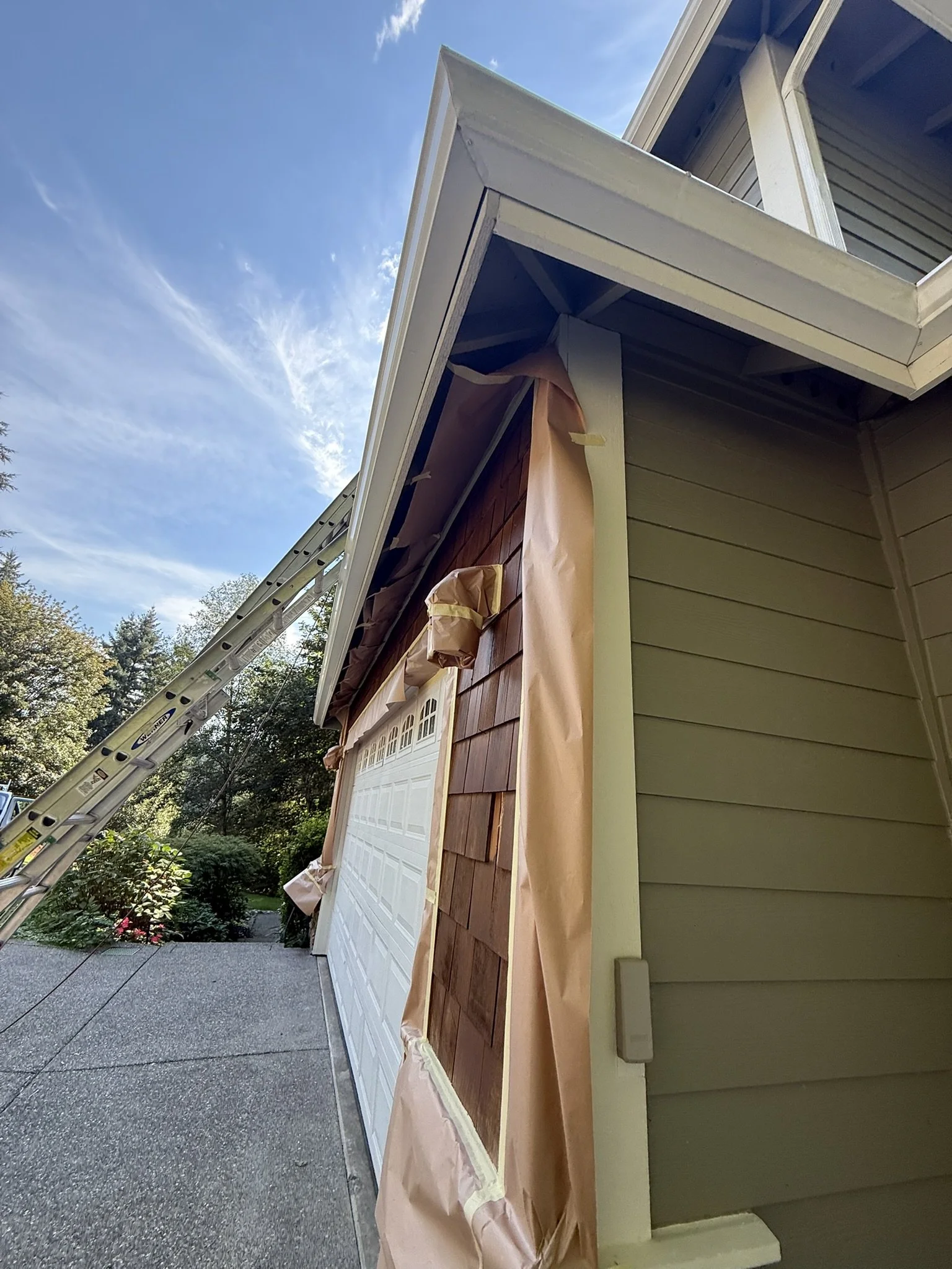 House exterior wall with papered and taped section for new siding or painting, ladder leaning against the house, partly cloudy sky, trees in background.