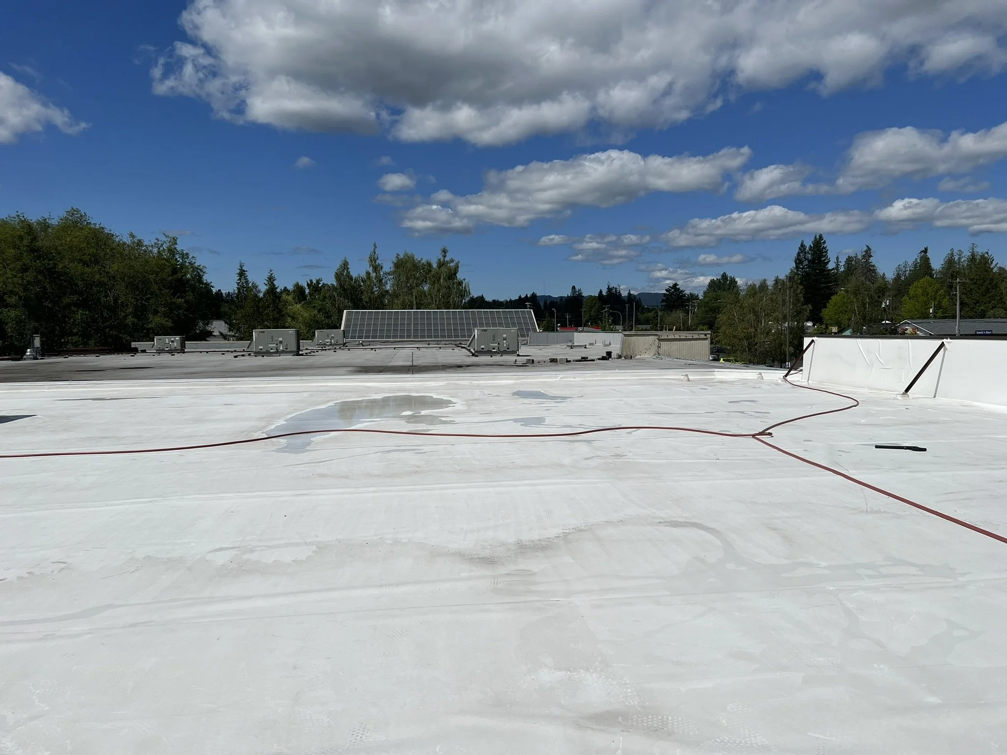 View of a building rooftop with a white reflective surface, a few puddles of water, and some red hoses or cables, under a partly cloudy blue sky with scattered clouds. Trees and buildings are visible in the background.
