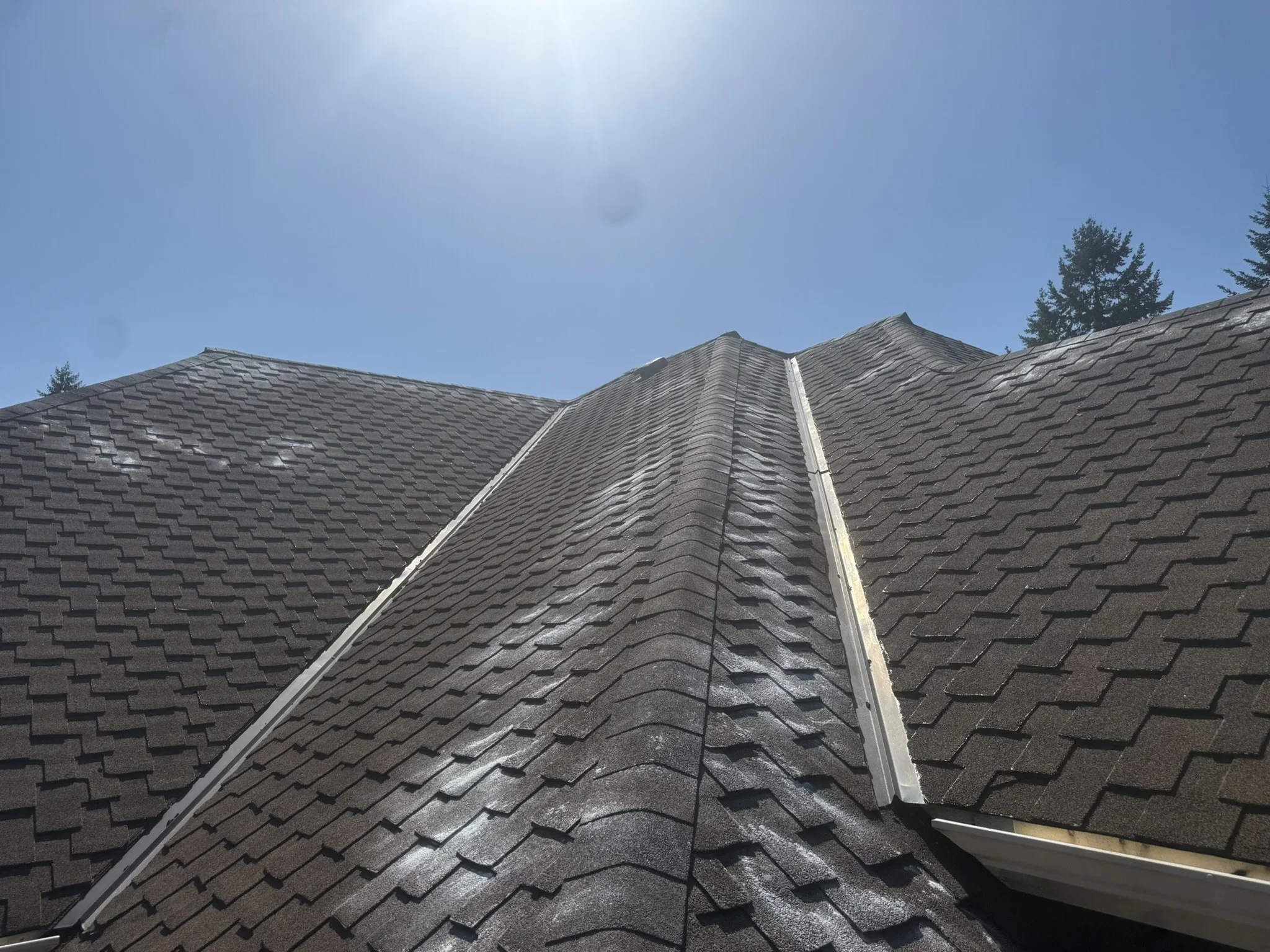 Close-up view of a sloped roof with brown shingles, with some white patches and a bright sun overhead in a clear blue sky.