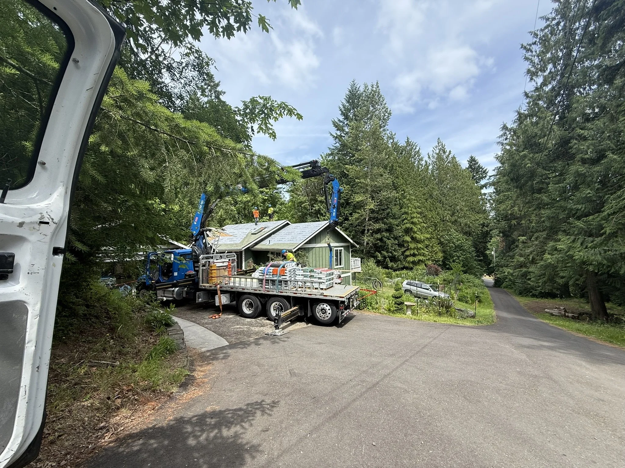 Construction workers on a roof using a crane truck, with a house amidst trees and a driveway leading away.