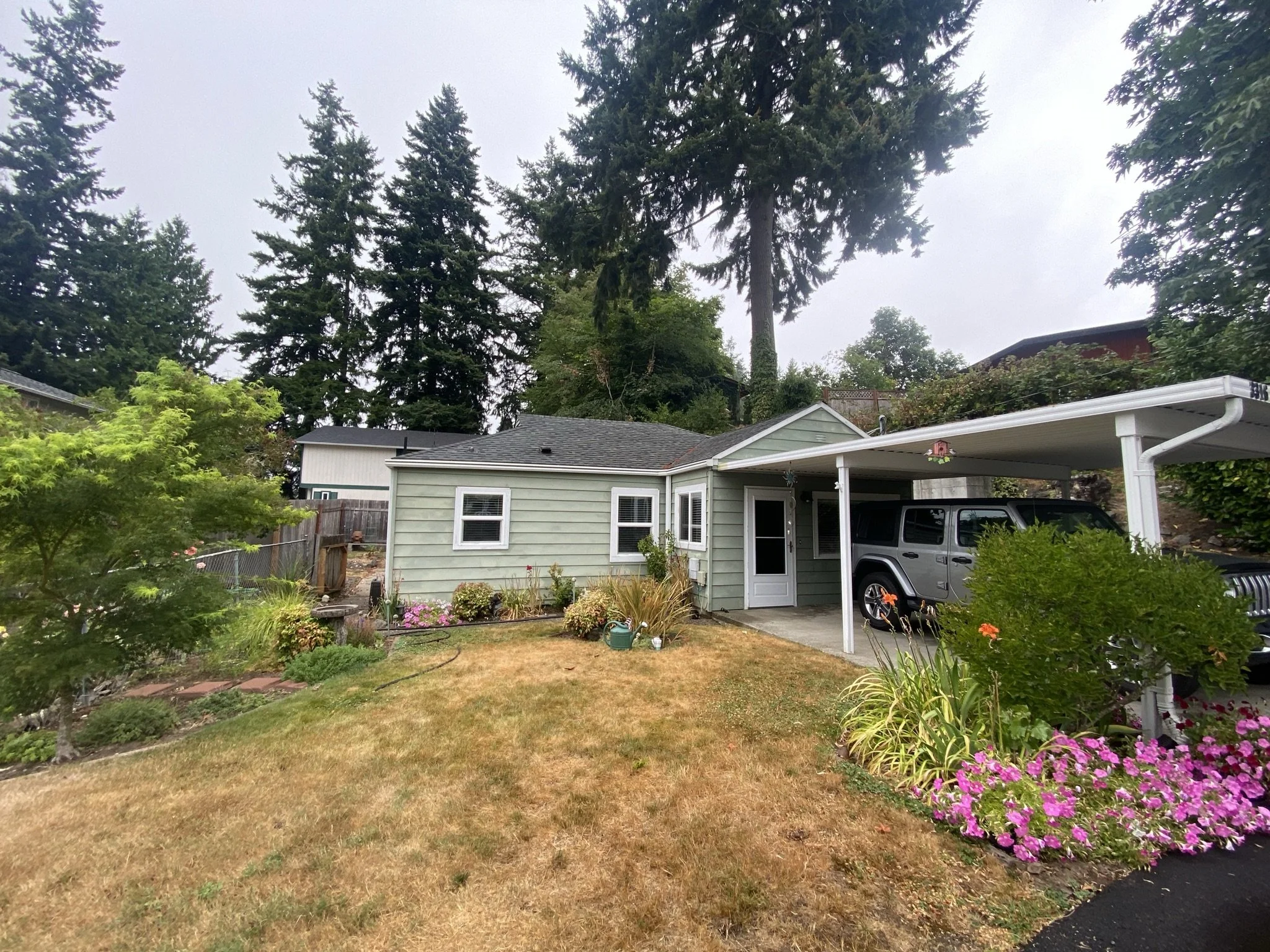 A suburban house with a carport, a gray Jeep, and a garden with pink flowers and greenery, tall trees in the background.