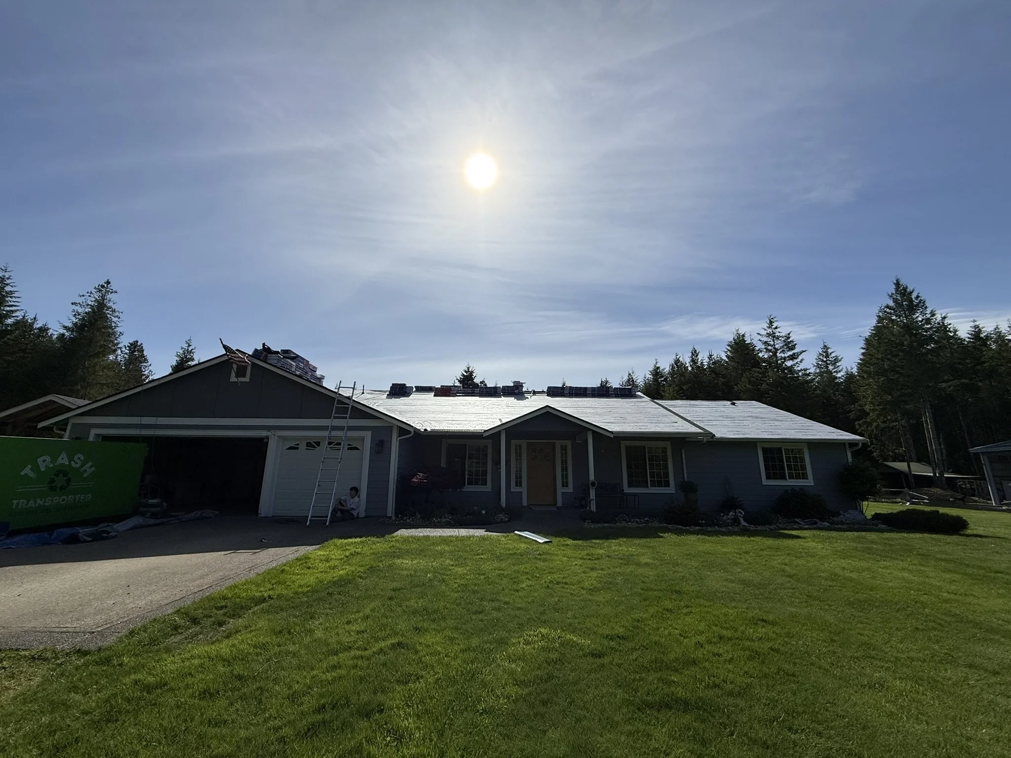 A house with a dark gray exterior and white trim, with solar panels being installed on the roof. The front yard has green grass, and there is a person sitting on the driveway. The sky is clear with the sun shining brightly.