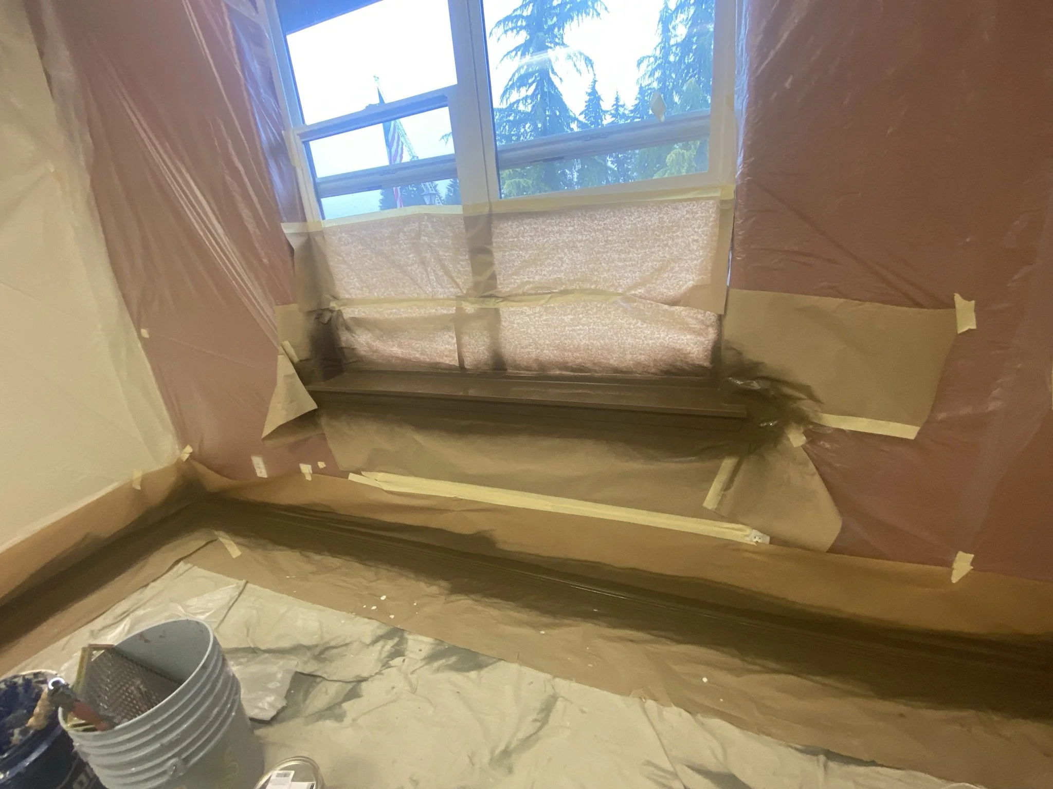 Room under renovation with plastic sheeting covering the walls and window, masking tape securing the plastic, and a bucket with painting supplies on the floor.