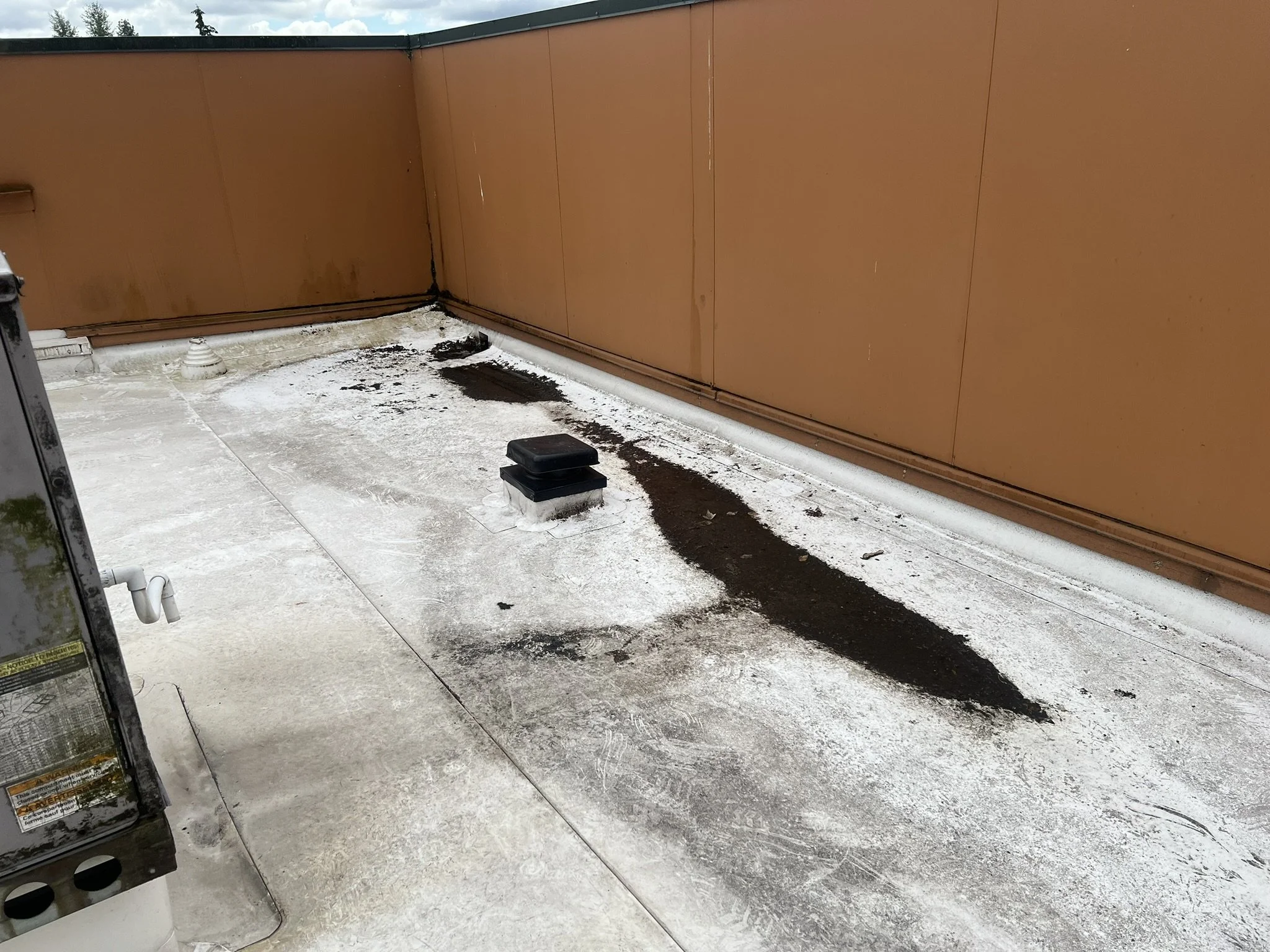 On a rooftop, there is a patch of dark dirt or asphalt on a white surface, with a brown wall in the background. Two black vents or covers are visible on the surface.