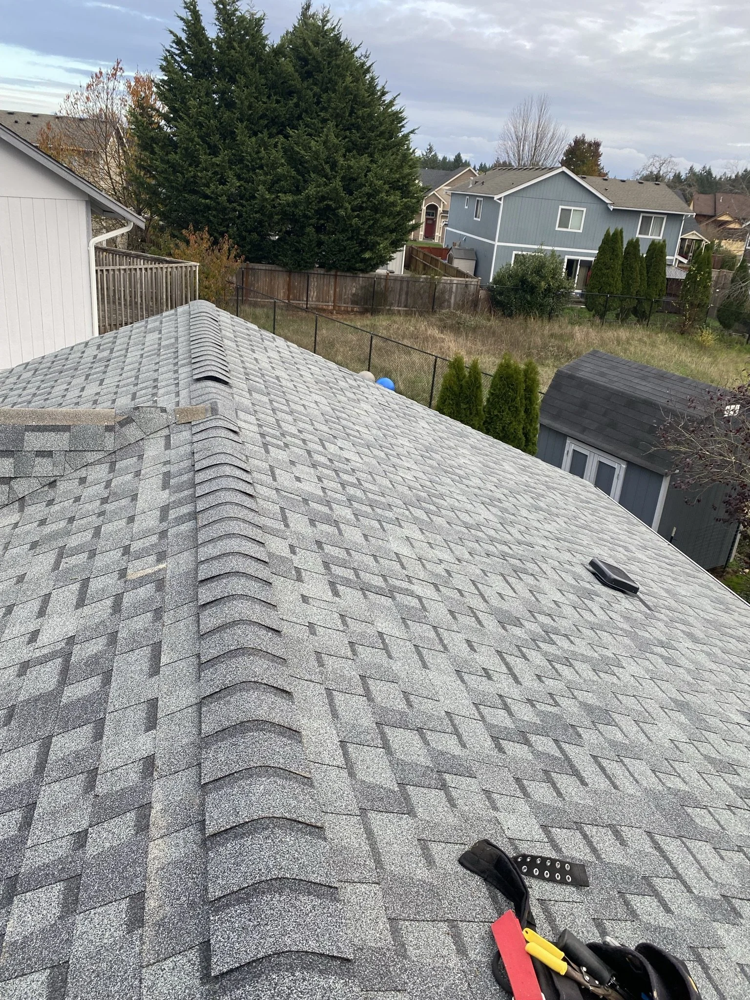 View of a gray shingled roof of a house with a roof vent and tools in the foreground, and neighboring houses, trees, and a fenced backyard in the background under a cloudy sky.