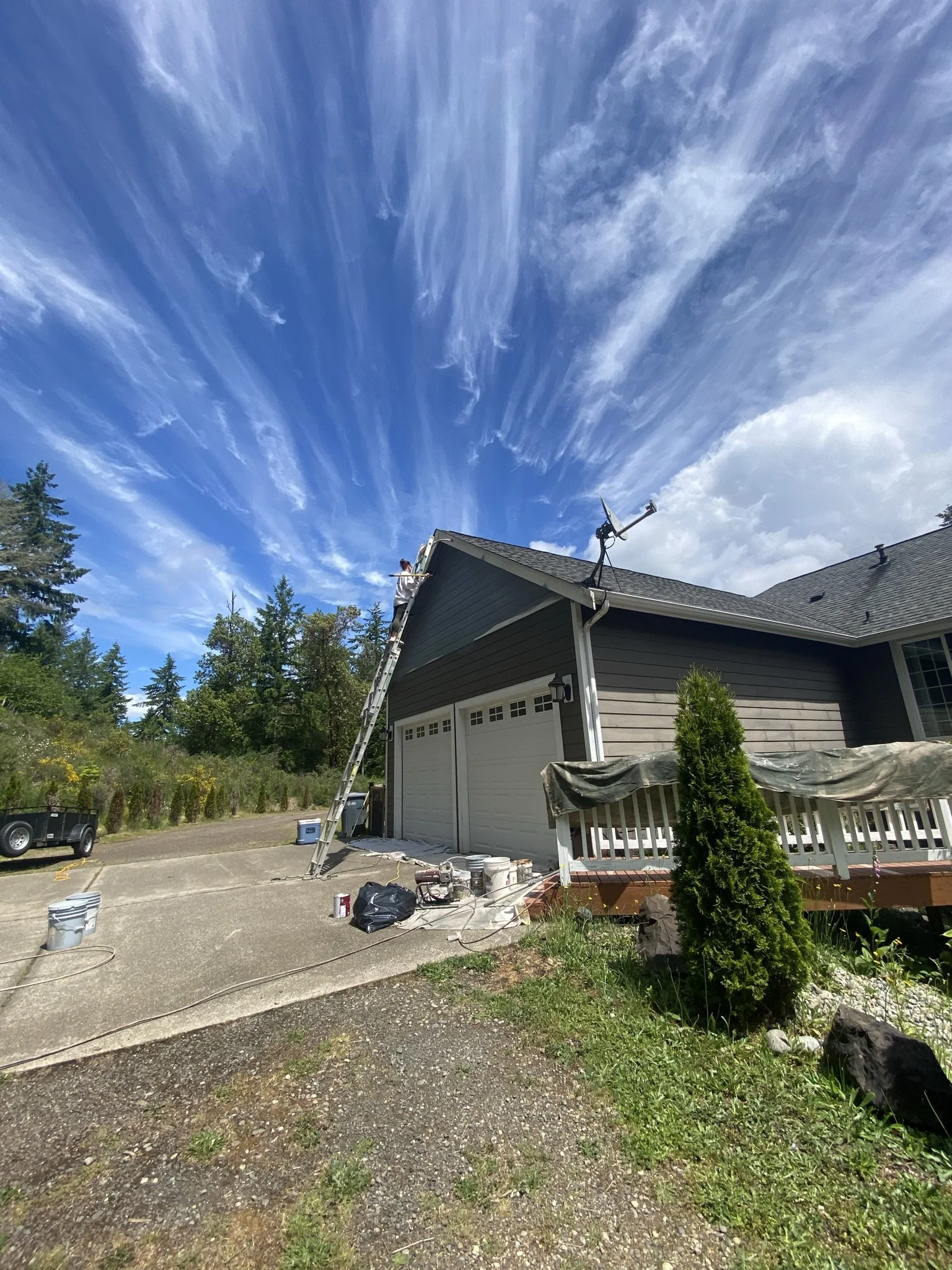 A house with a ladder leaning against the roof and a person working on the gutter or roof area. The sky is partly cloudy with streaks of clouds, and there are trees and a garden in the background.