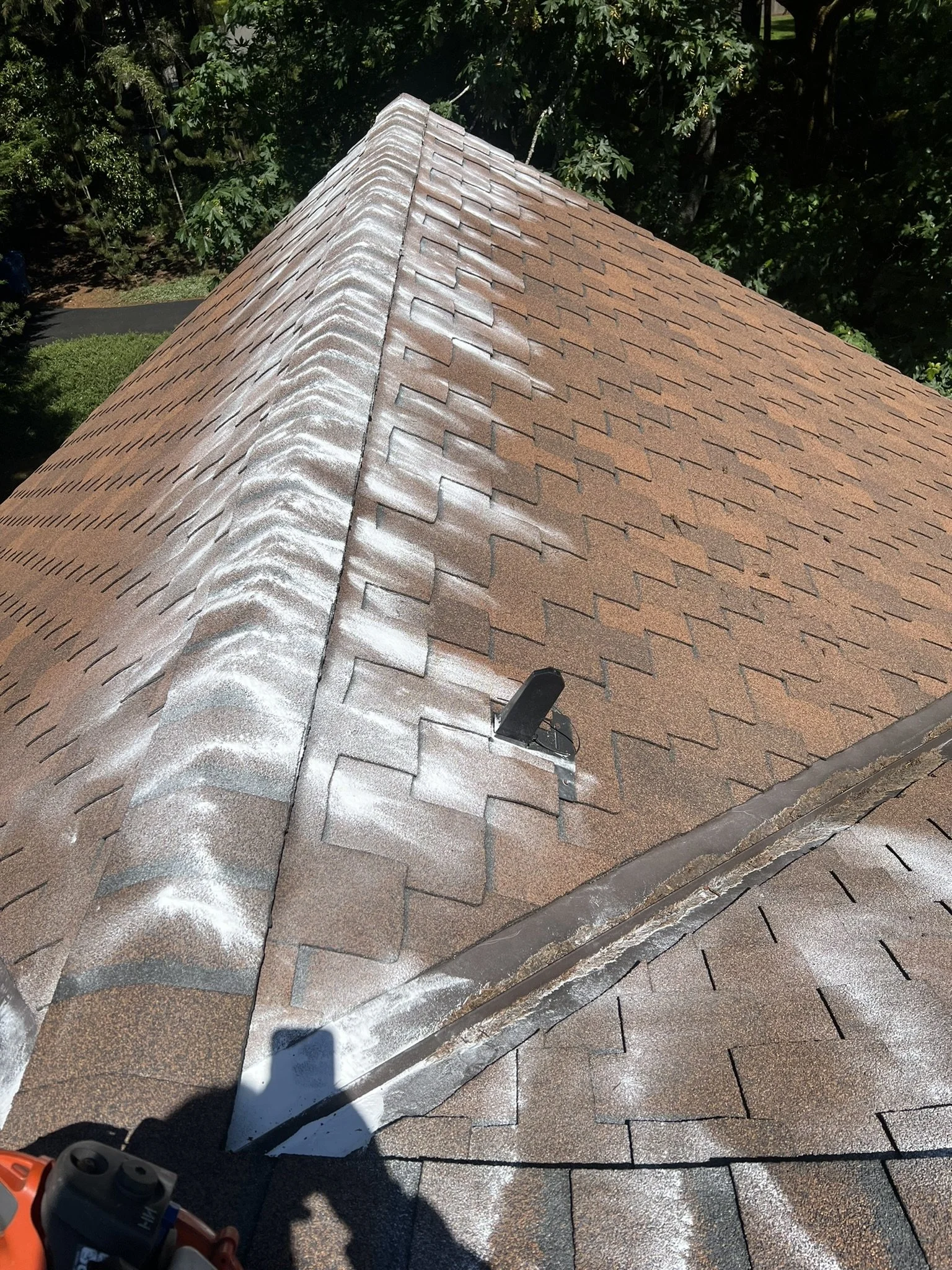 From a high angle, a roof with brown asphalt shingles is shown, featuring a central ridge. Some shingles are coated with white residue, possibly indicating cleaning or treatment. A black vent pipe is installed on the roof, and a person's hand holding