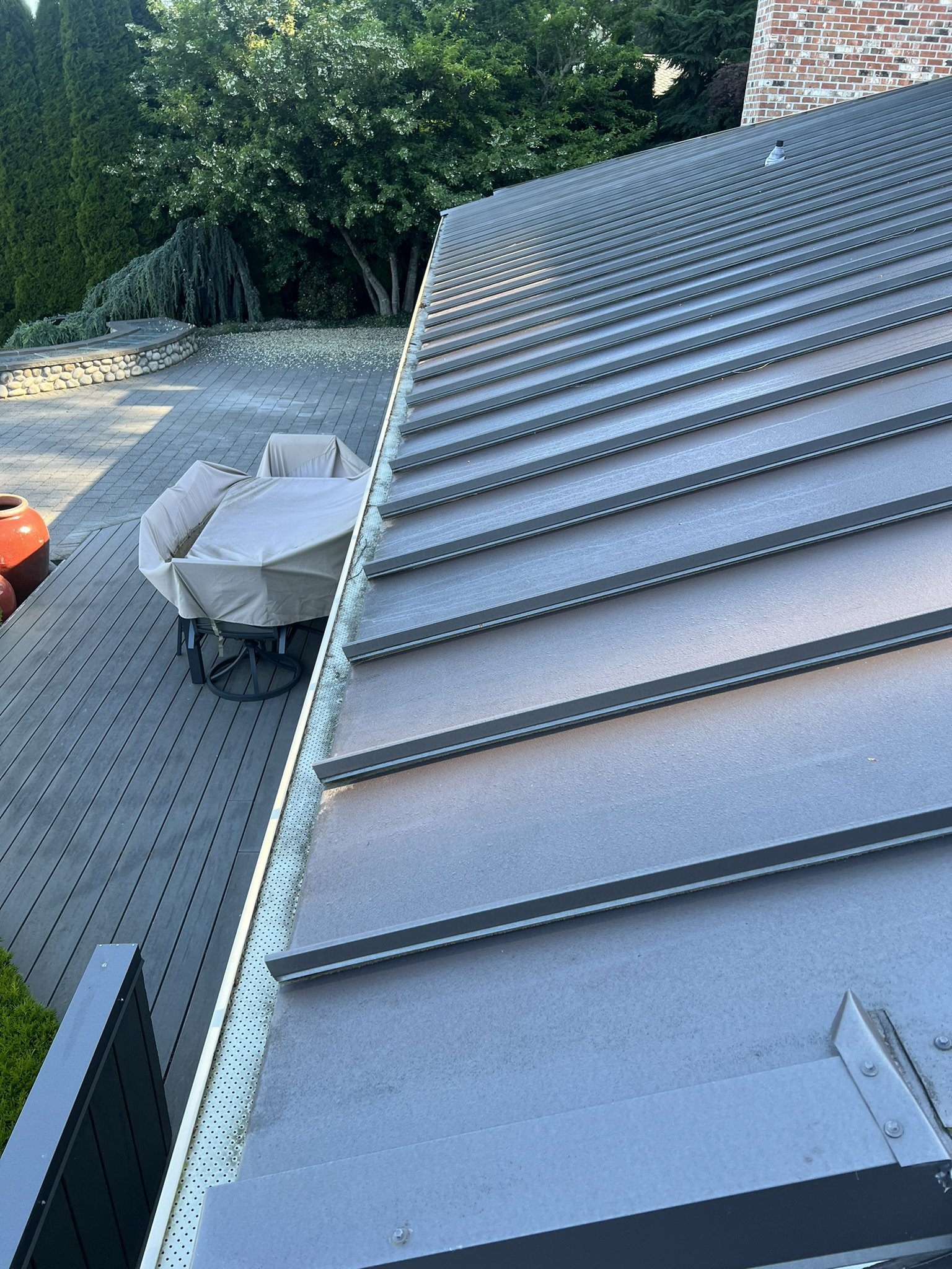 Side view of a metal roof with standing seam panels, adjacent to a deck area with outdoor furniture, surrounded by green trees and landscaping.