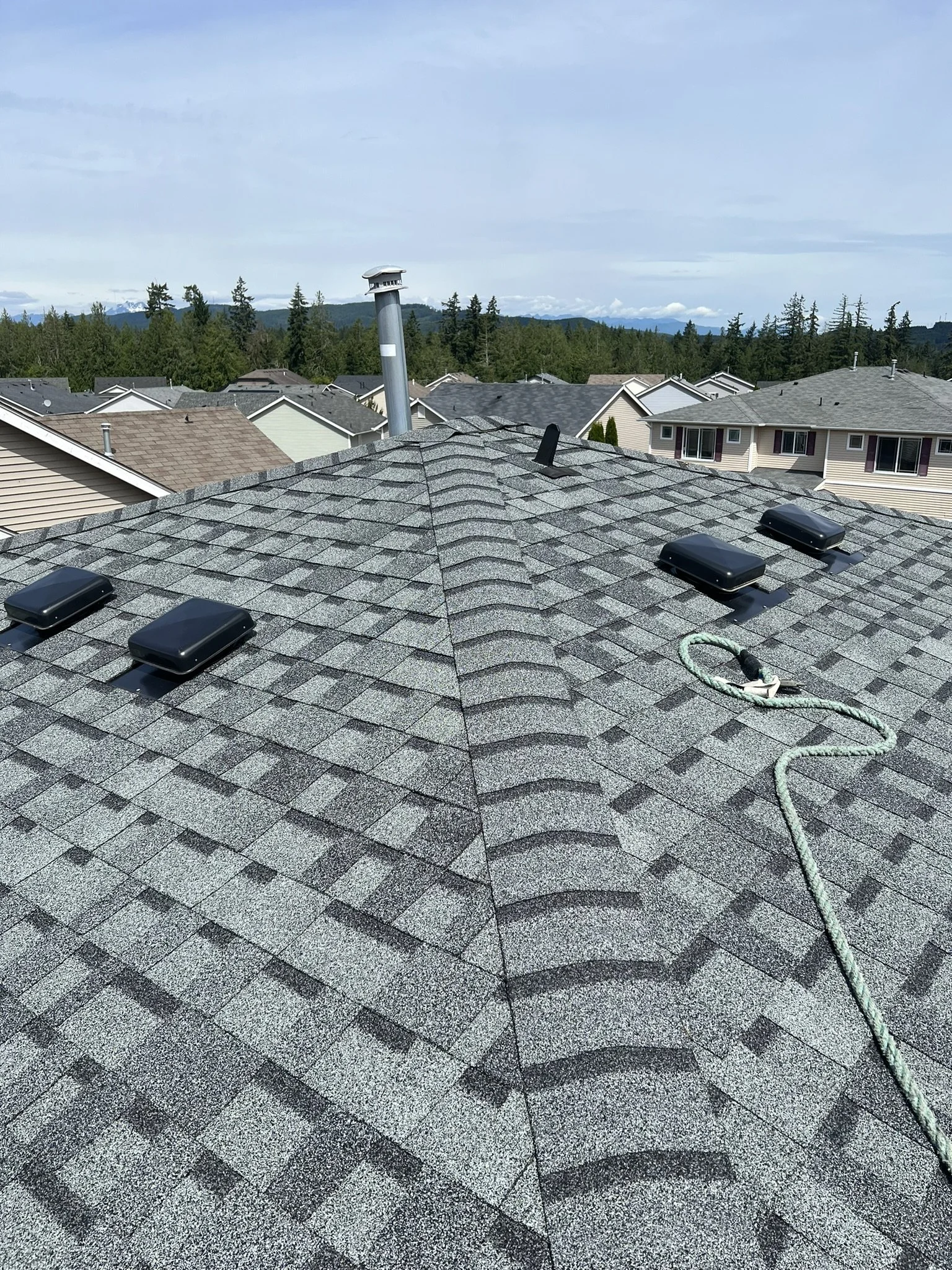 View of a residential rooftop with asphalt shingles, four black vent covers, a chimney pipe, and a climbing rope. Neighboring houses, trees, and mountains are visible in the background under a partly cloudy sky.
