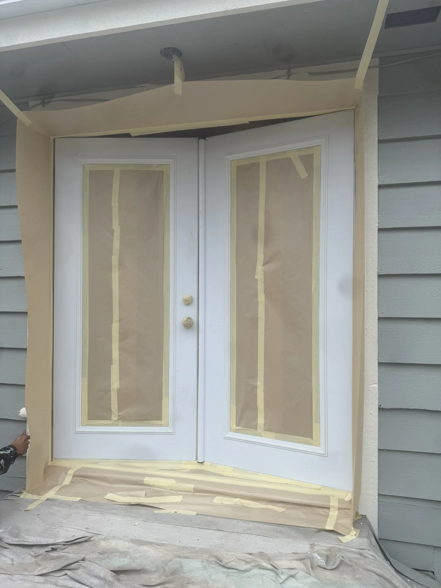 Front door with glass panels covered by masking paper and tape for painting, surrounded by beige painter's paper, in a house exterior with gray siding.
