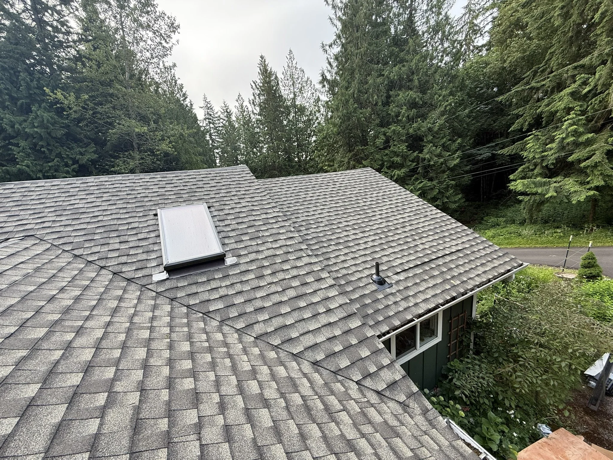 View of a house roof with grey shingles, a solar skylight, and a vent, surrounded by green trees and a road in the background.