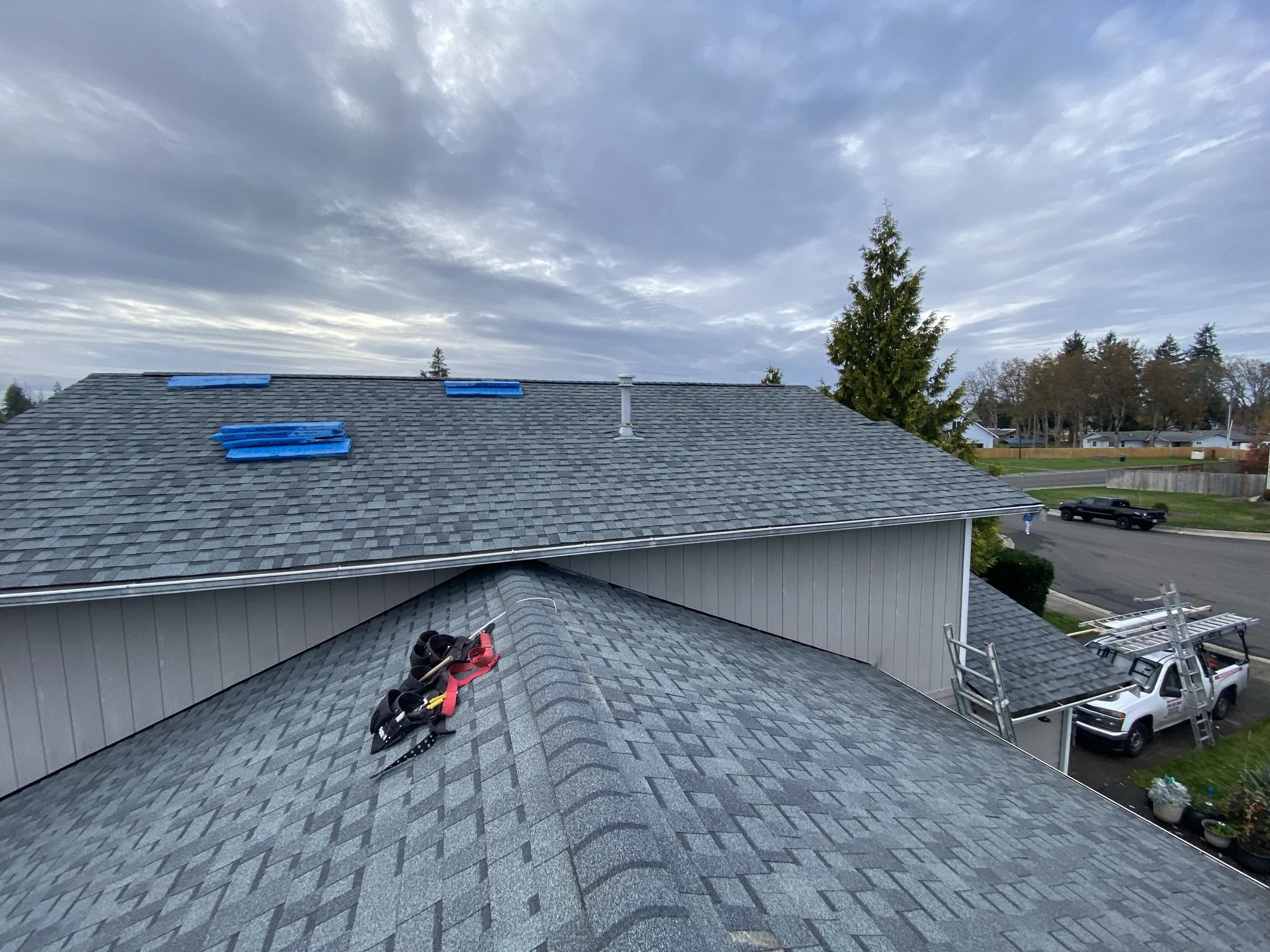 Residential roof with shingles, with tools such as harnesses and ladders, likely for roofing work, under a cloudy sky.
