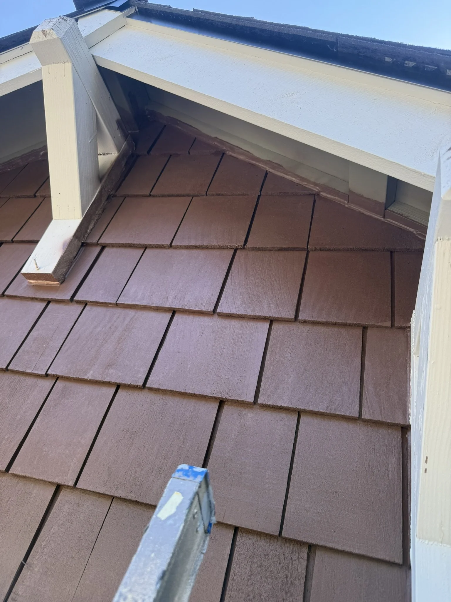Roof with brown shingles and a small dormer window under a blue sky.