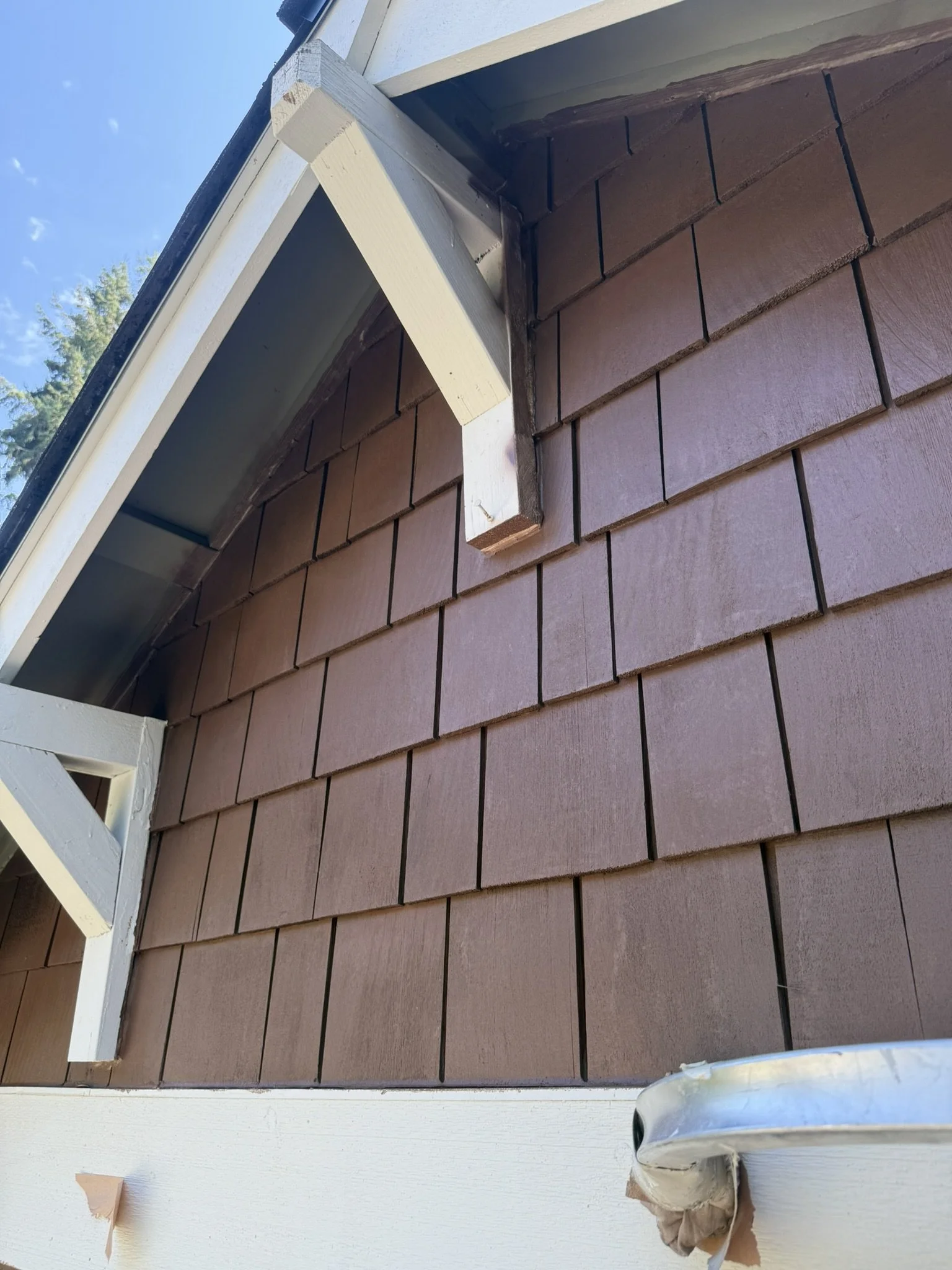 Close-up of the corner of a house wall with brown shingle siding, white trim, and part of the roof, showing a small section of the sky and trees in the background.