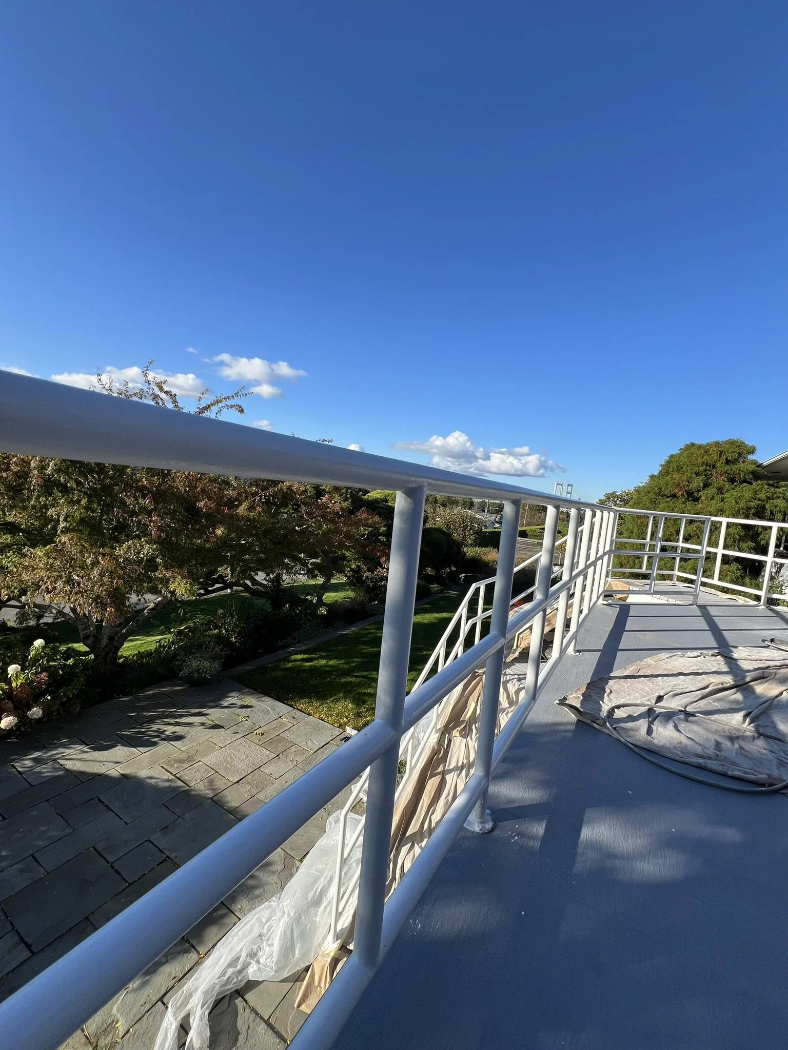 View from a balcony with white metal railing overlooking a garden with trees, bushes, and a stone pathway under a blue sky with white clouds.